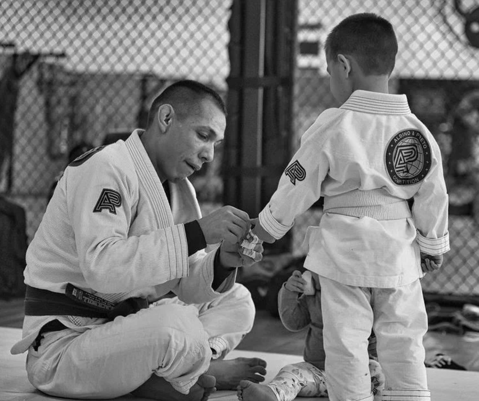 A Brazilian Jiu-Jitsu instructor kneeling on the mat and helping a young student with her uniform in a martial arts gym.