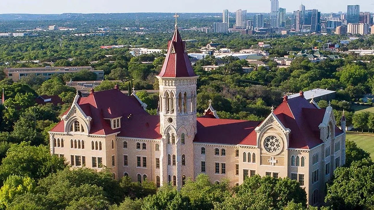 A large historic church with a red roof, white walls, and a central tower, surrounded by green trees and set against a city skyline in the background.