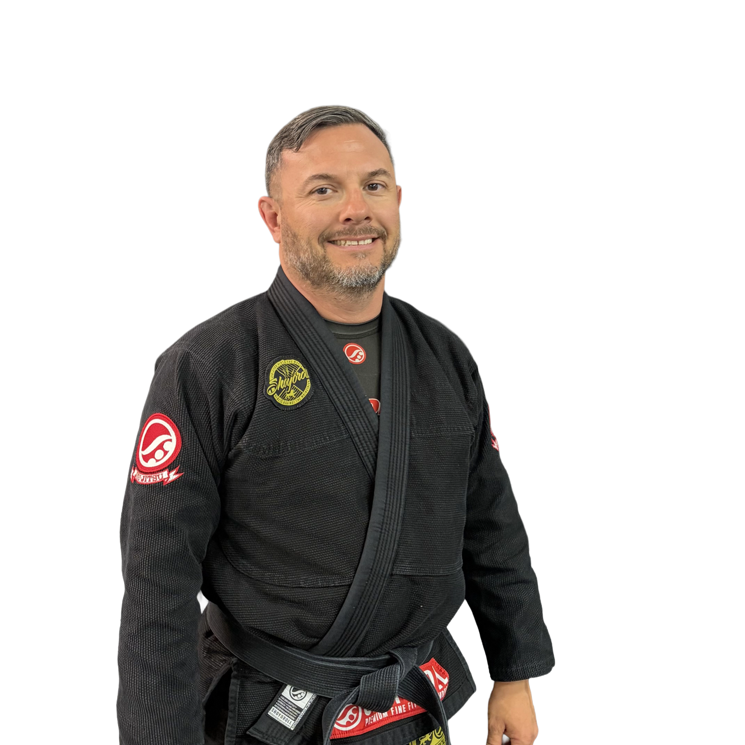 Man in a black Brazilian Jiu-Jitsu gi with patches, smiling, standing against a white background.