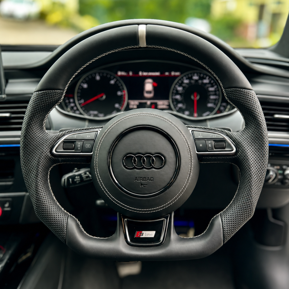 Close-up view of an Audi car steering wheel with the Audi logo, digital dashboard with speedometer and tachometer in the background, and various control buttons on the wheel.