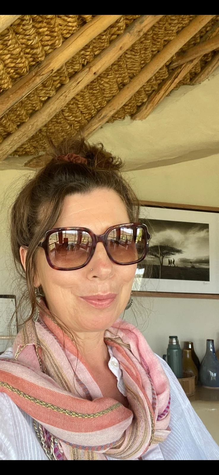 Woman with brown hair in a messy bun, wearing large sunglasses and a pink patterned scarf, smiling indoors with artwork and decorative bottles in the background.