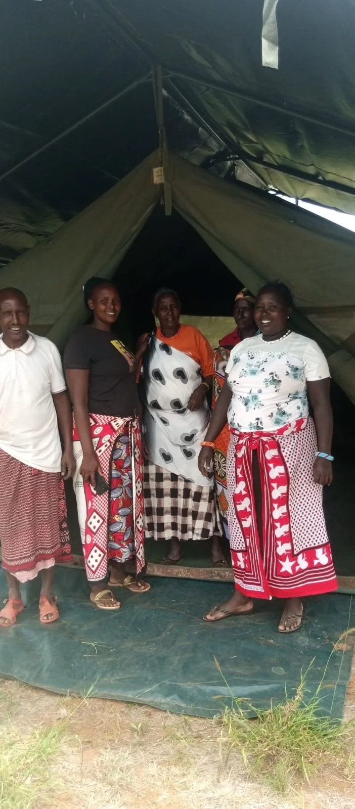 Group of five women and one man standing inside a large green tent, smiling, wearing colorful traditional and casual clothing.