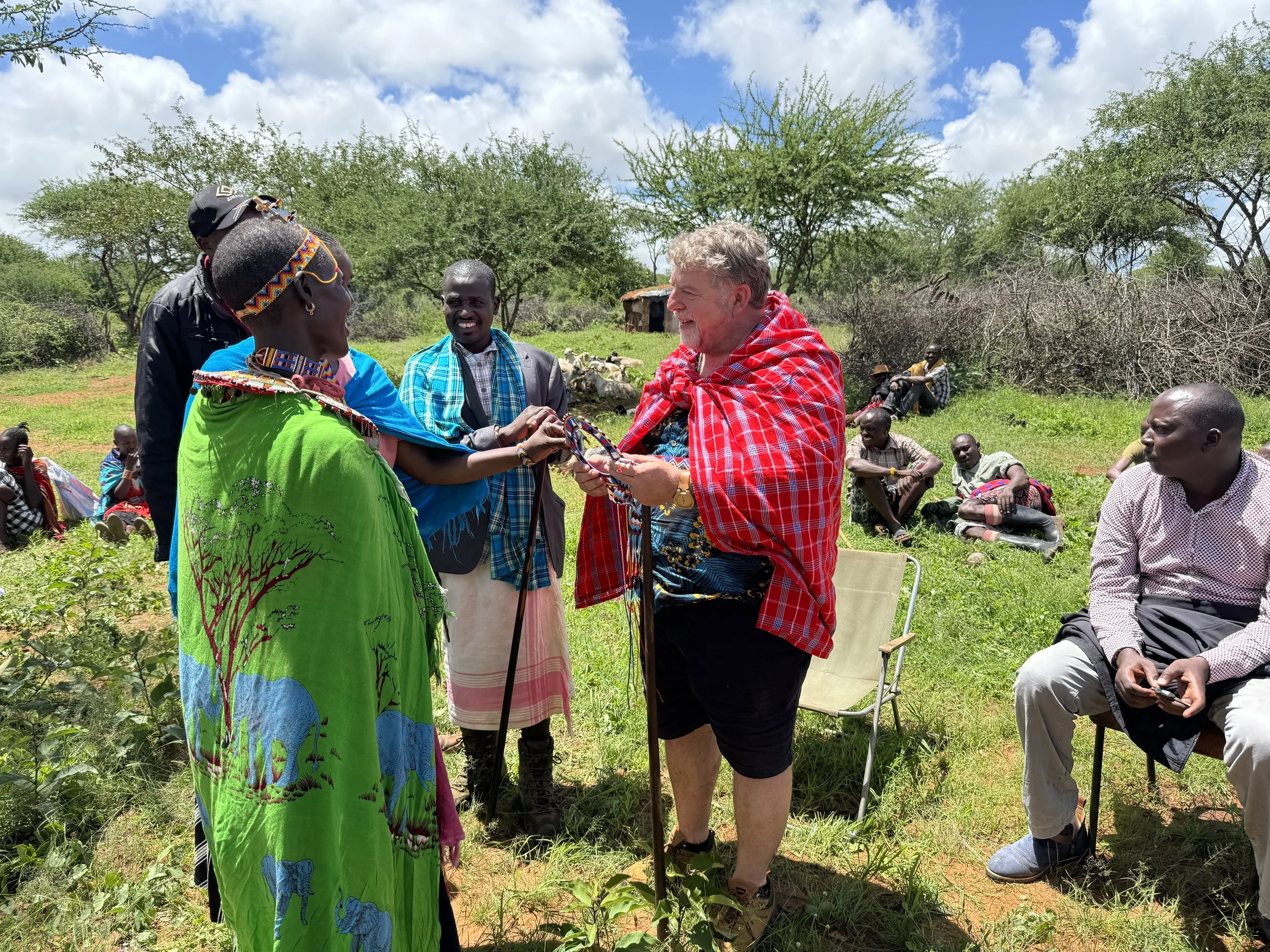 A group of people outdoors on a sunny day, with a woman in traditional clothing holding hands with a man in a plaid shawl, while others sit or stand nearby, surrounded by trees and a small hut in the background.