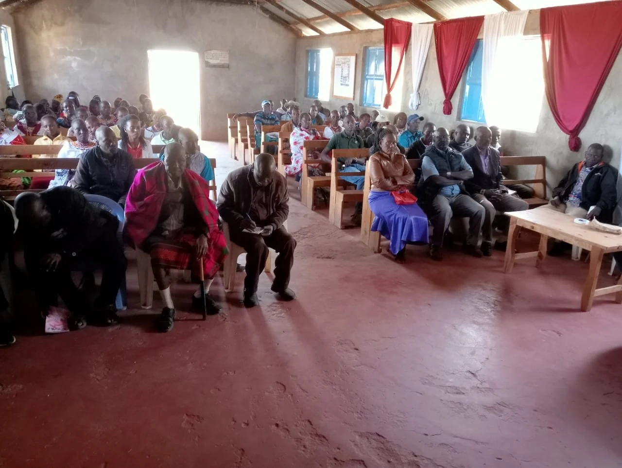 A group of people seated inside a simple room with a red dirt floor, blue windows, and red and white curtains, attending a gathering or meeting.