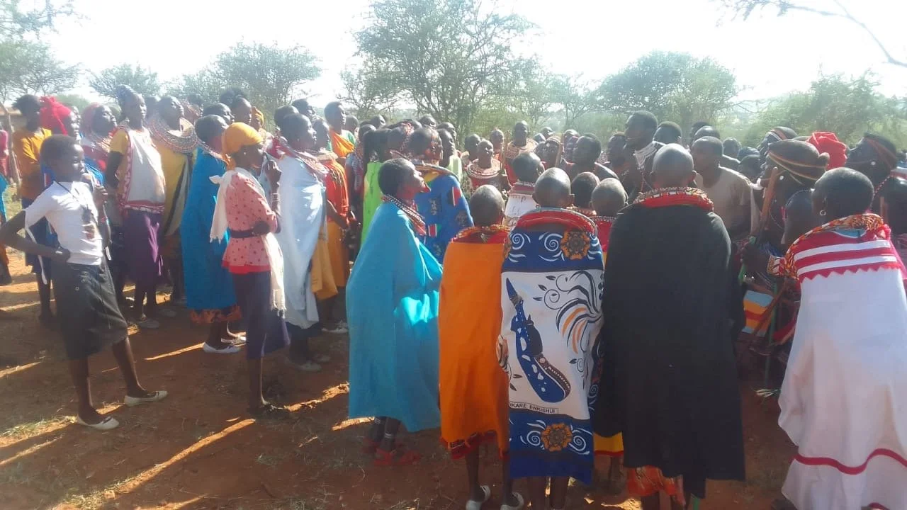 A large group of people dressed in colorful traditional clothing, gathered outdoors on a dirt ground, with trees in the background, likely participating in a cultural event or gathering.
