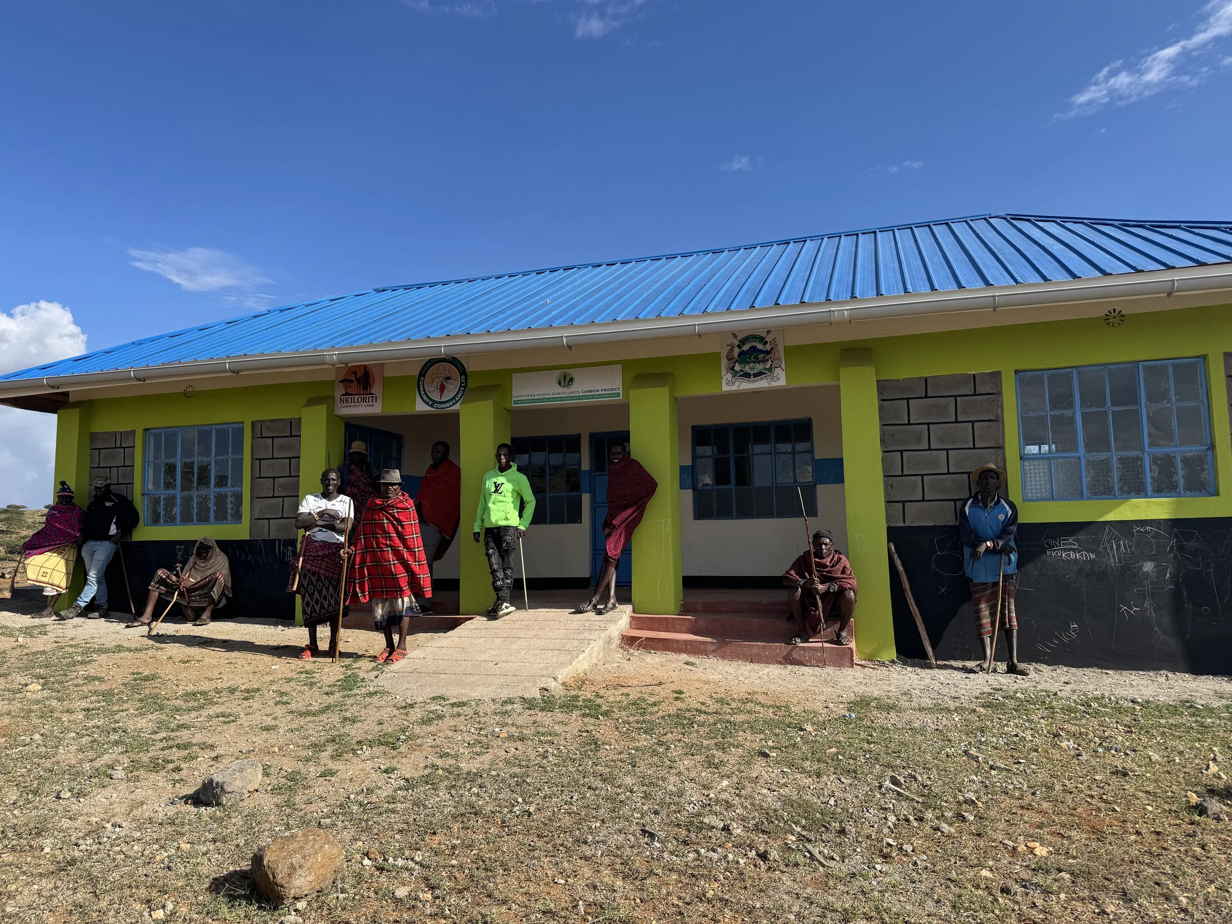 A group of people gathered in front of a building with lime green walls and a blue roof. The building has several windows and signs on the front. People are standing and sitting, some wearing traditional clothing, under a clear blue sky.