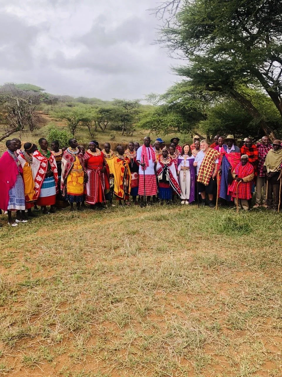 A group of Maasai and other indigenous people in traditional clothing gathers outdoors on a grassy area with trees and hills in the background.