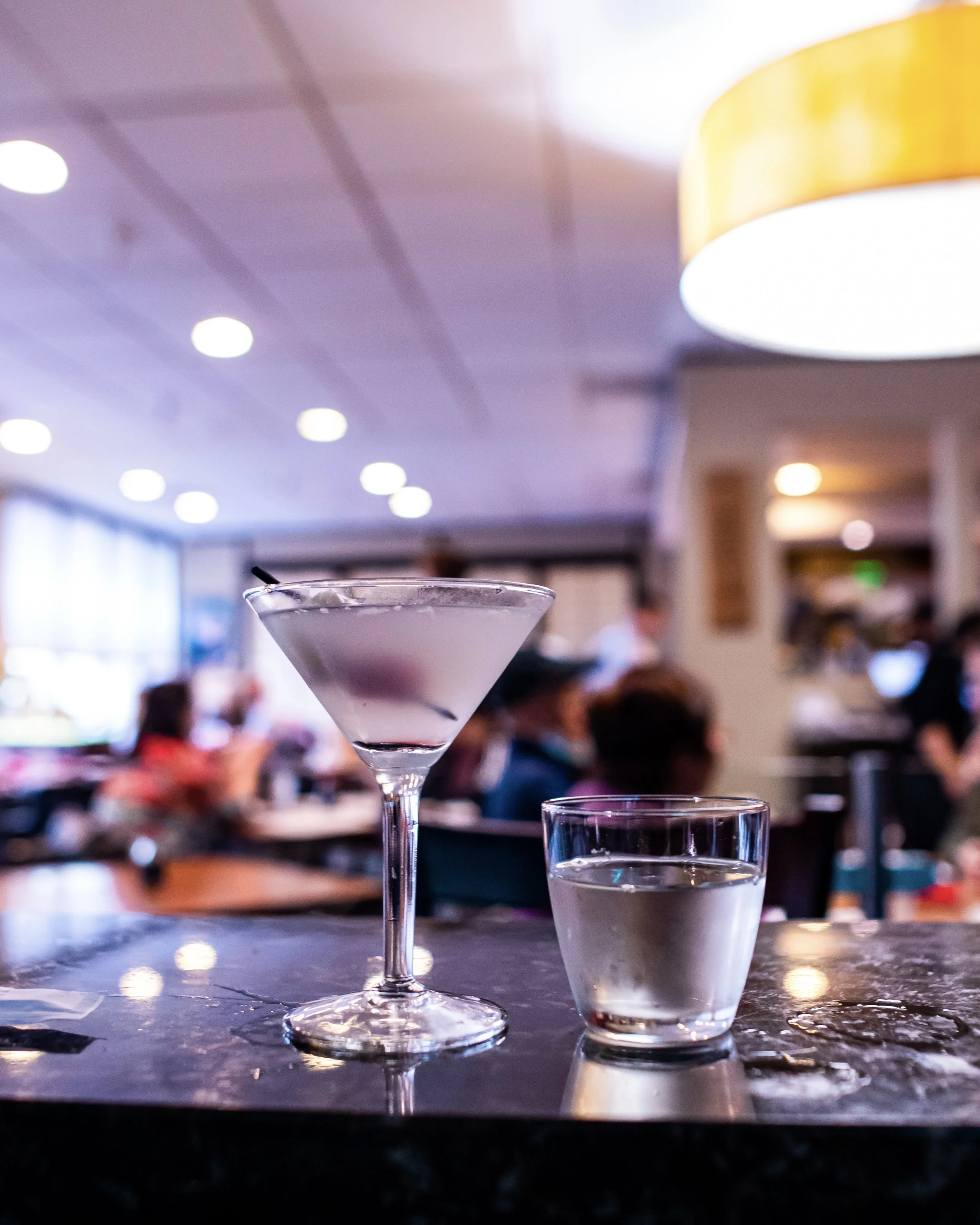 A pink cocktail in a martini glass and a glass of water on a dark table in a busy restaurant.