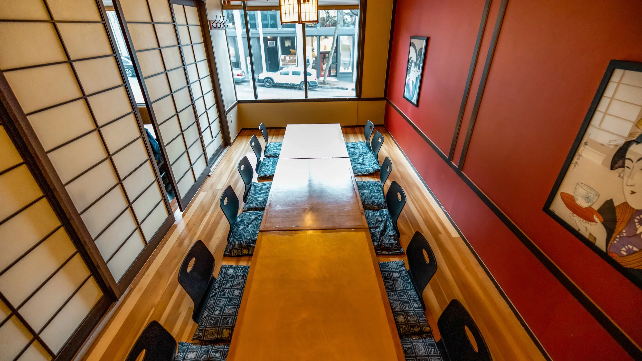 A rectangular dining table with a light wood finish, surrounded by nine black chairs with patterned cushions on a wooden floor inside a Japanese-style restaurant, with shoji screens on one side and framed artwork on the red wall.