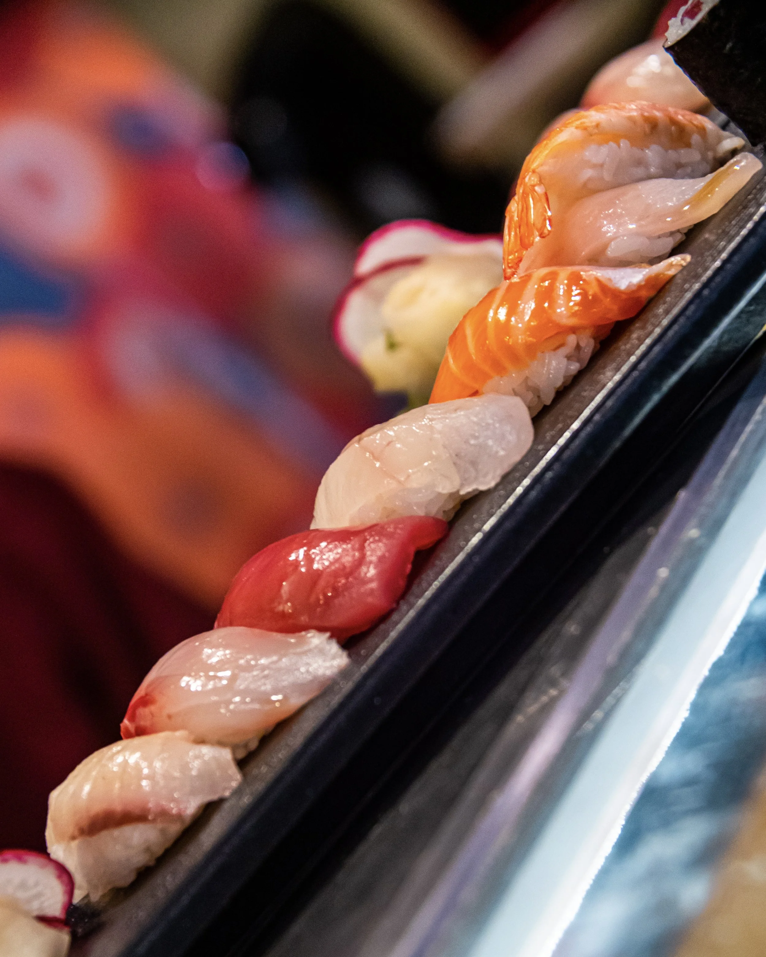 Close-up of assorted sushi pieces on a black tray, including salmon, shrimp, and white fish, with colorful radish garnishes.