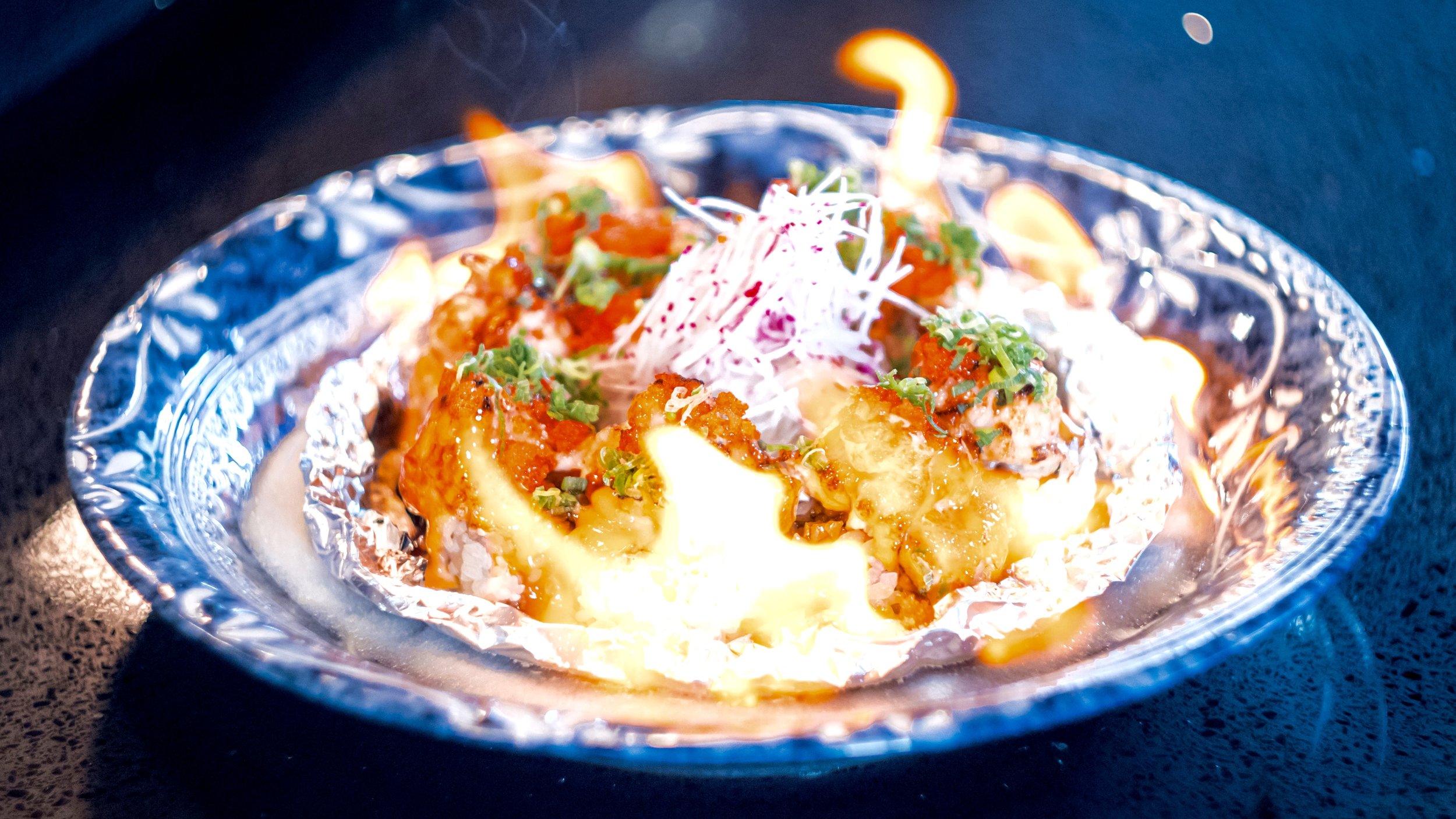 Baked Mexican street corn in a small foil tray garnished with green herbs, served with shredded cheese and sauce.