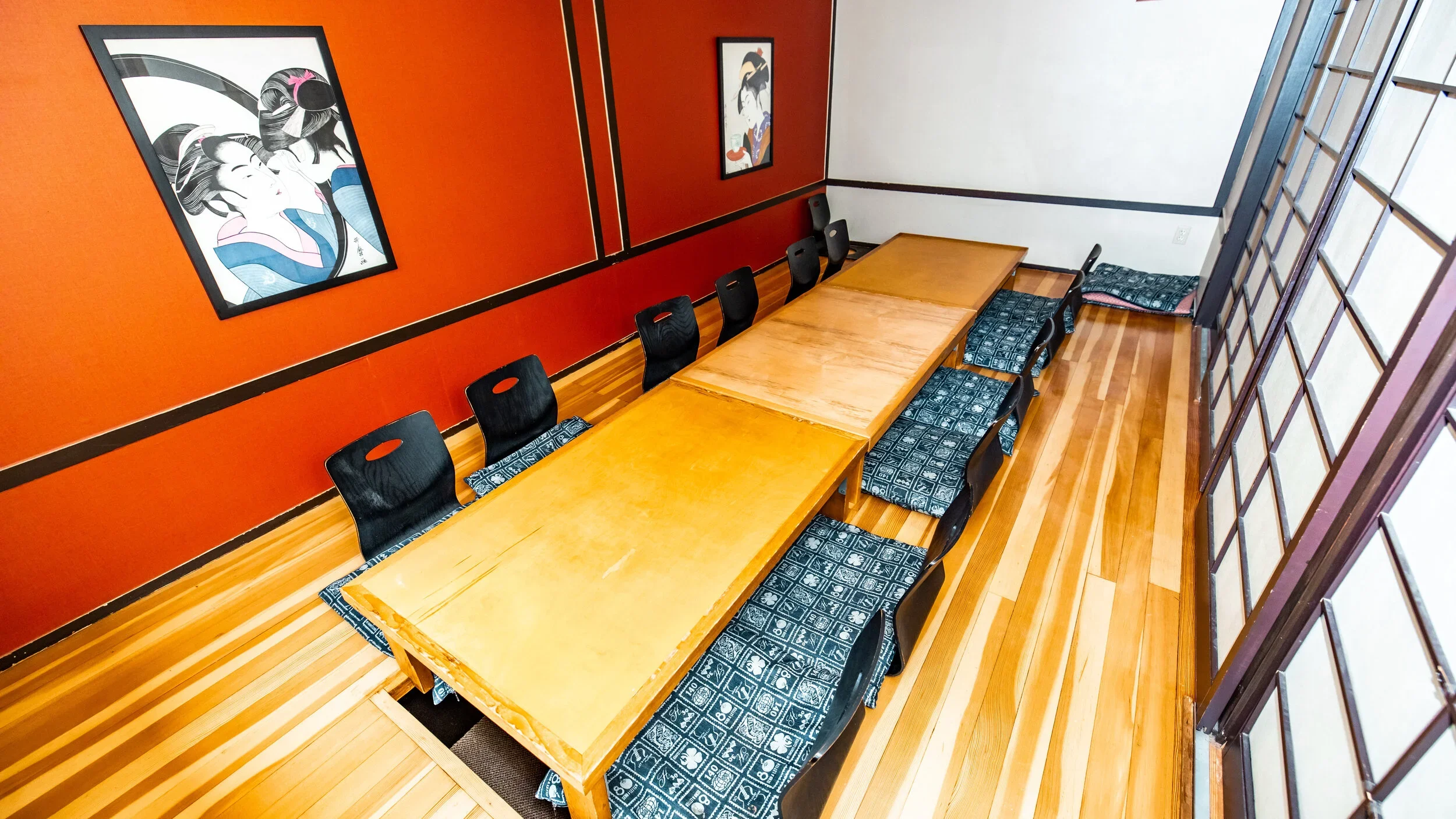 A Japanese-style dining room with a long wooden table, black chairs, traditional tatami mats on the floor, two colorful manga-style posters on a red wall, and shoji-style sliding paper doors.