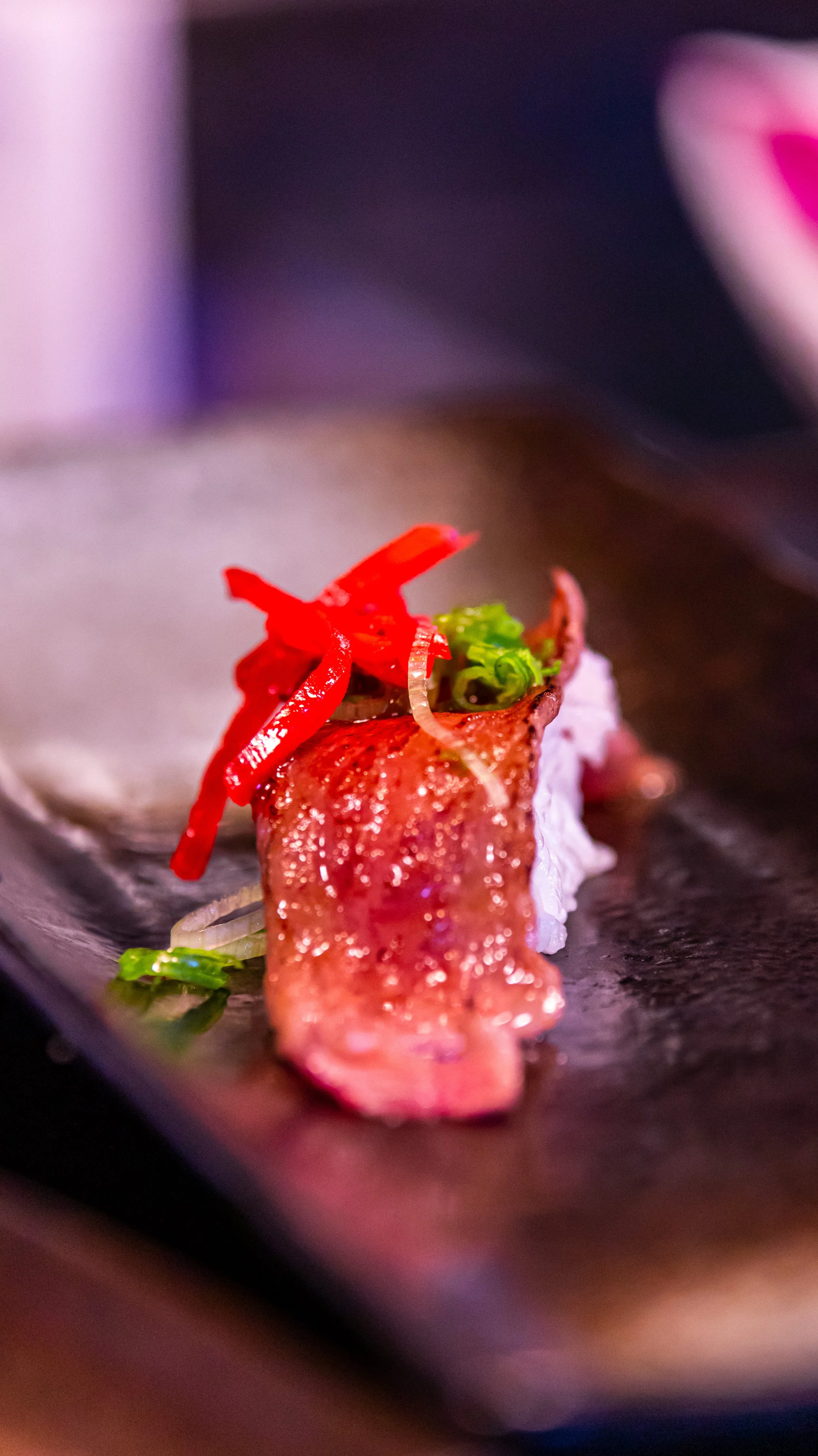 Close-up of a piece of sushi topped with red sliced chili peppers and green onions, on a black plate.