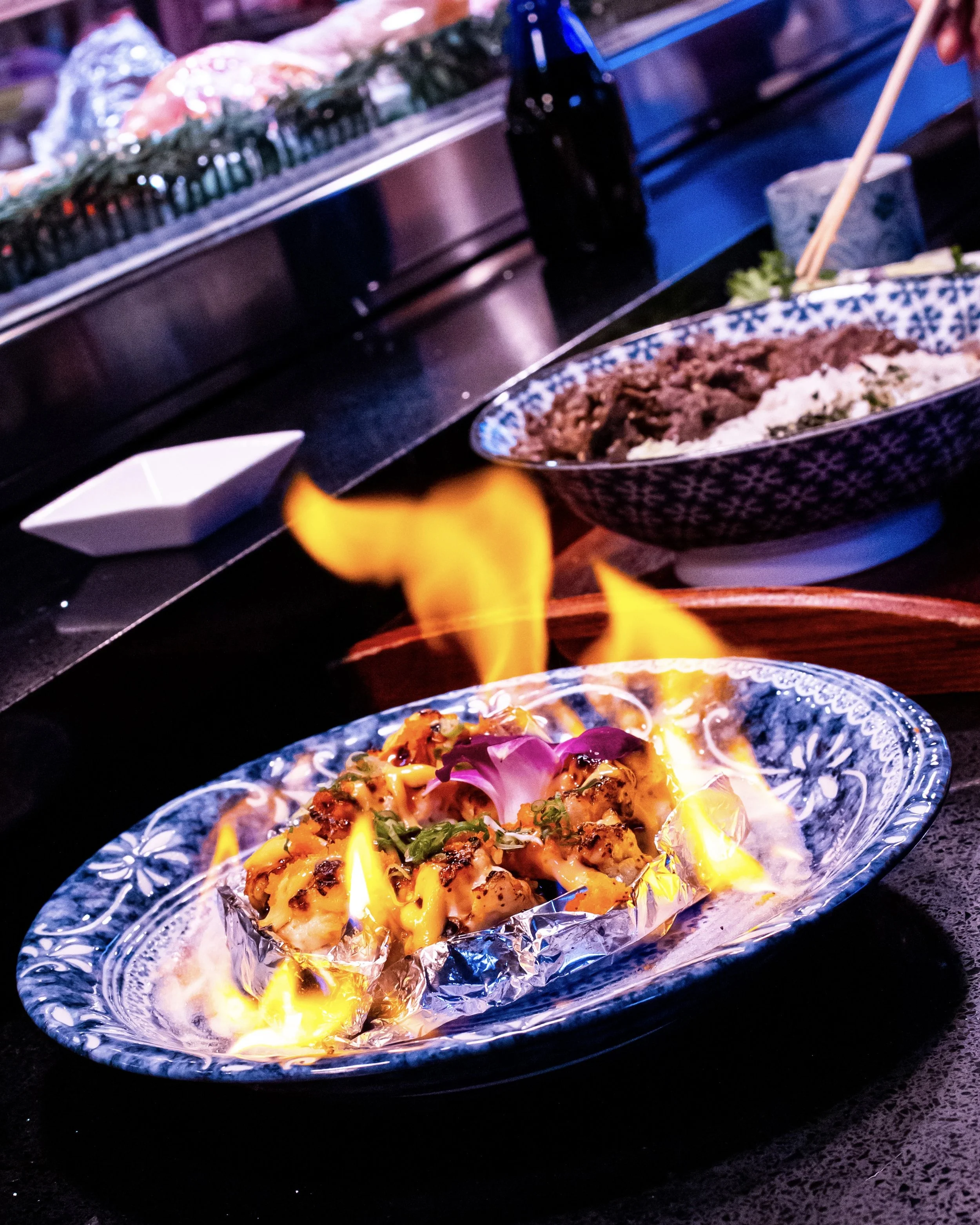 Food being flambéed on a decorative plate at a restaurant with dishes in the background.