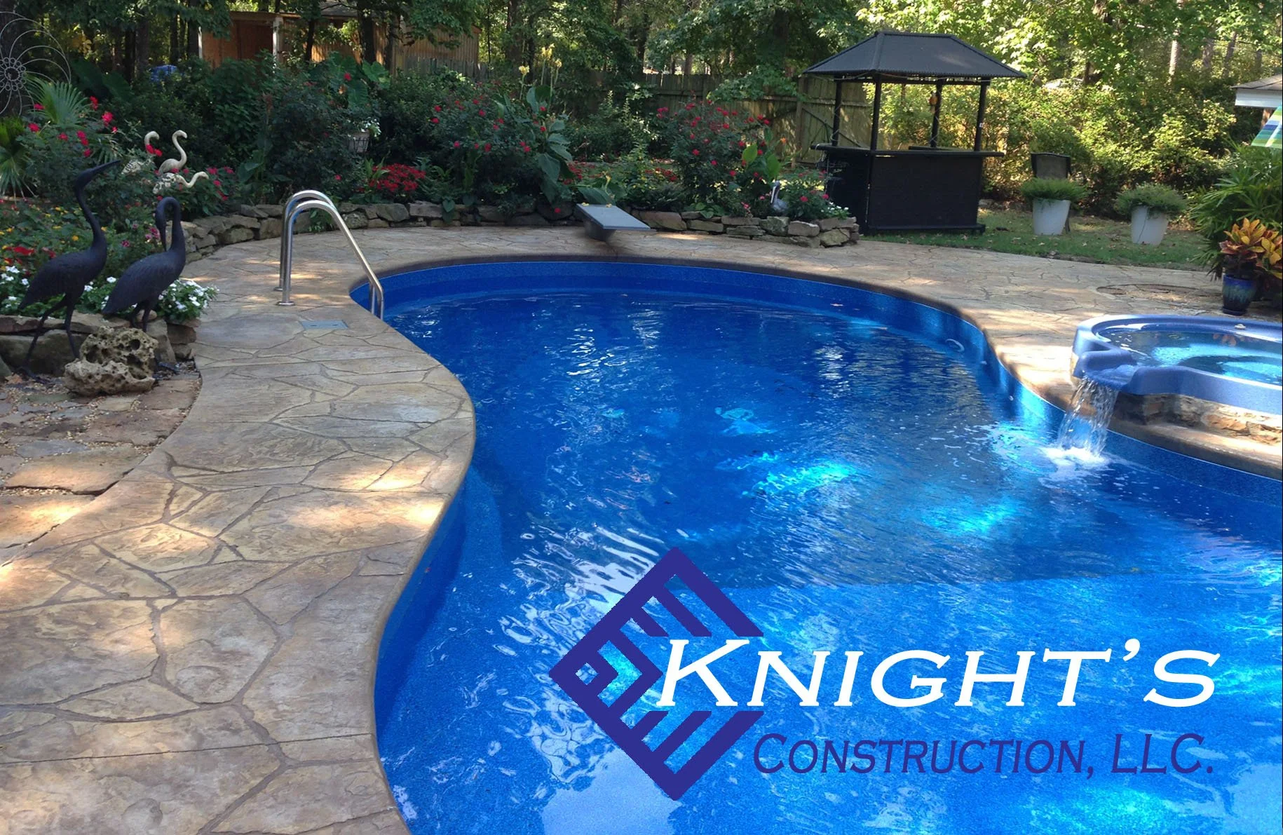 a backyard swimming pool with a small waterfall feature, surrounded by a stone deck, decorative metal heron statues, and lush garden plants, including flowers and shrubs, with a wooded area and a gazebo in the background.