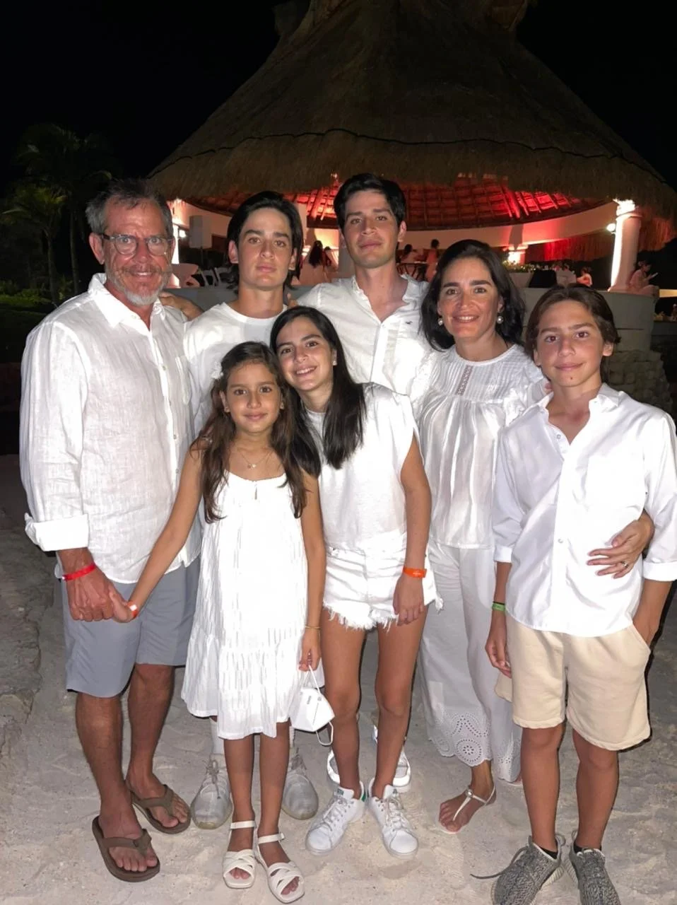 Familia de ocho personas en una celebración nocturna en la playa, con fondo de palapa, vestidos de blanco, sonriendo y disfrutando juntos.