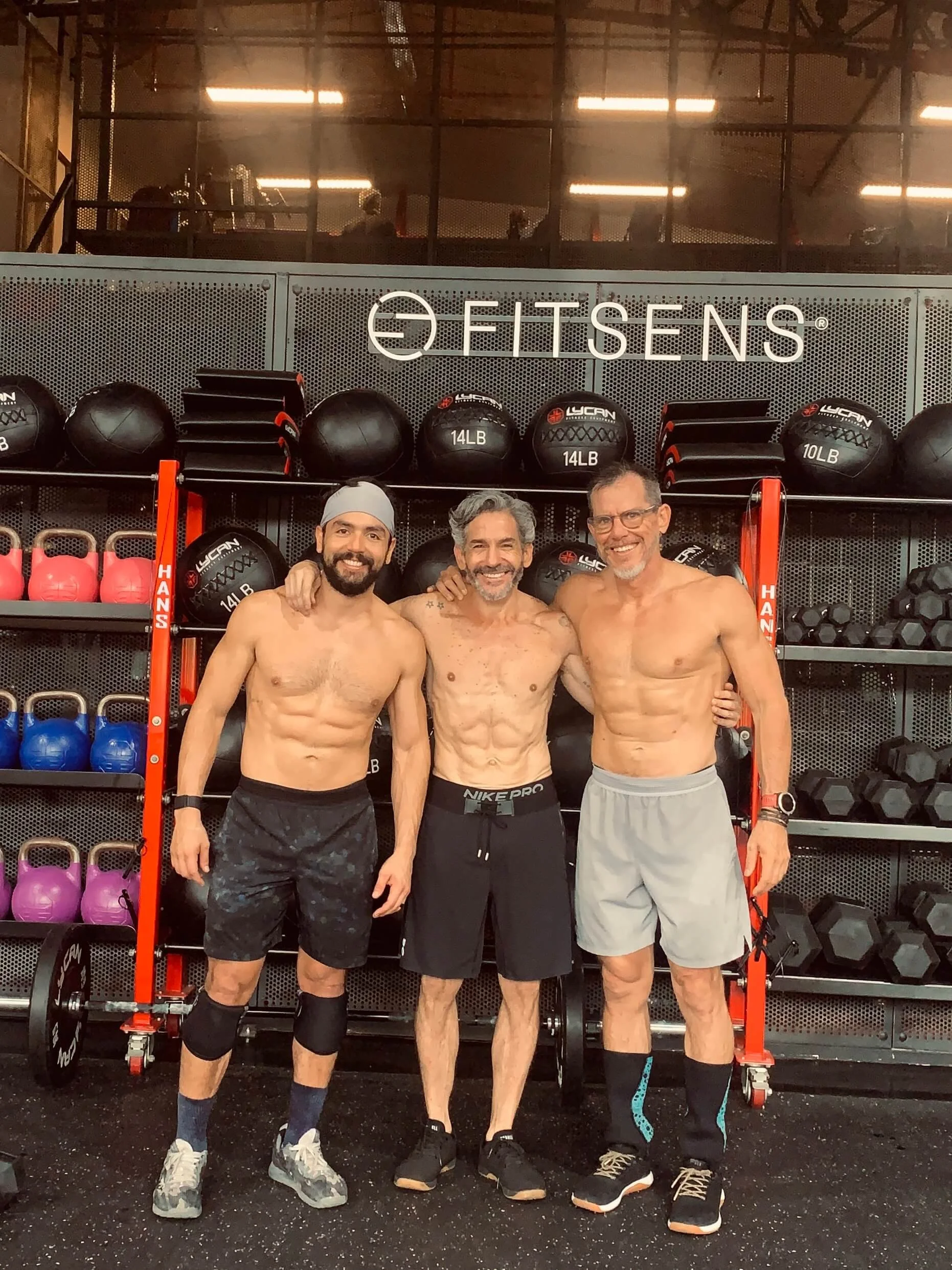 Tres hombres musculosos sin camiseta en un gimnasio, sonriendo y posando juntos.