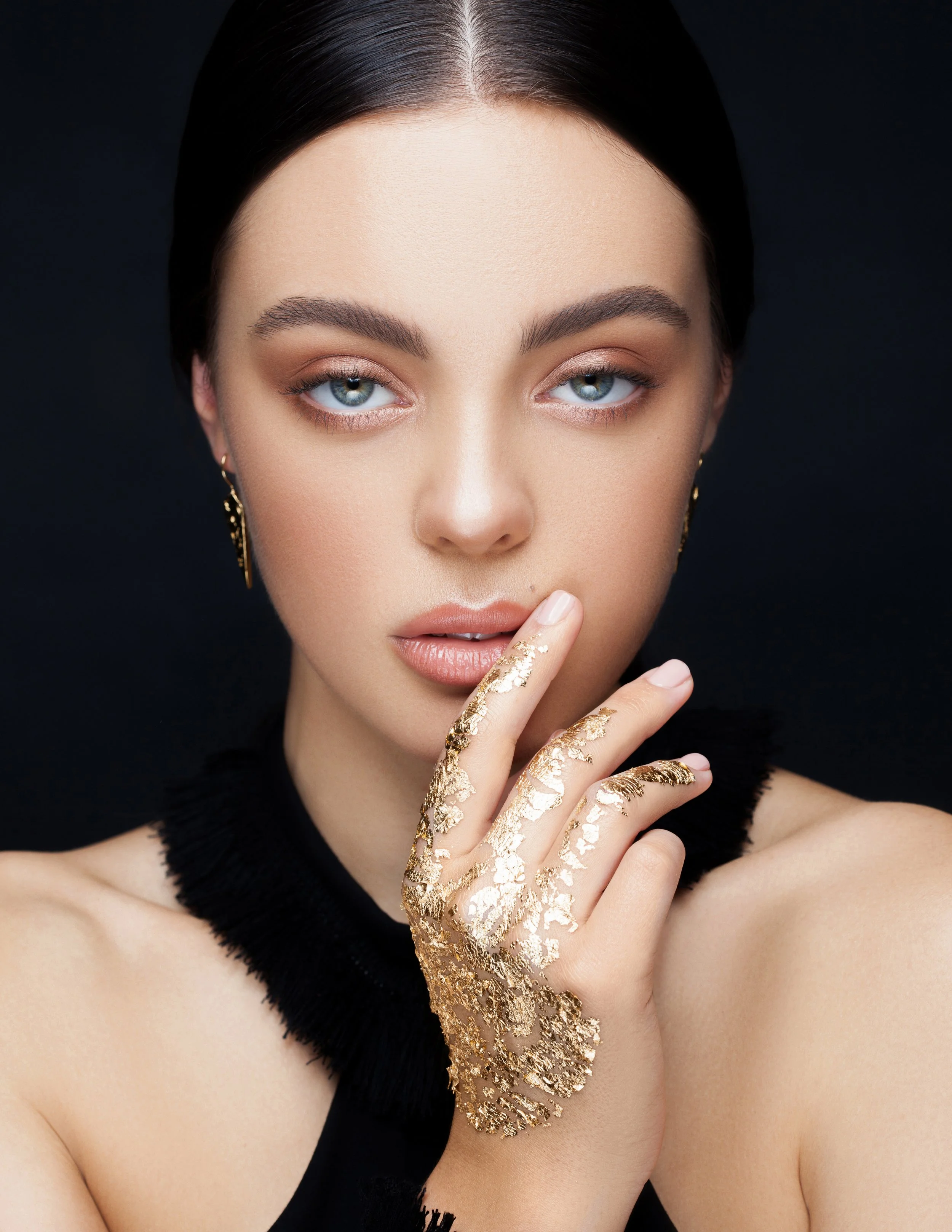 A close-up portrait of a woman with blue eyes and dark hair, wearing gold earrings, a black top, and with her hand covered in gold foil decoration near her face against a black background.