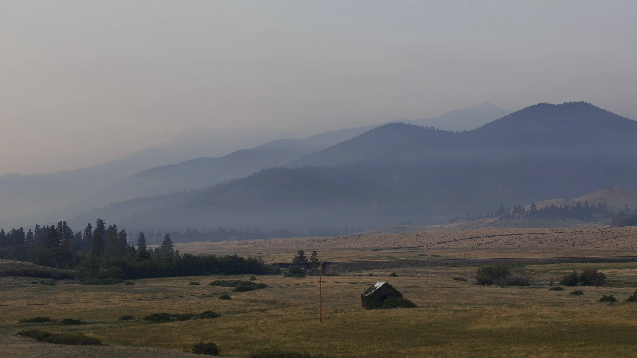 A scenic landscape featuring rolling hills and mountains with mist or fog, a small barn, and a power line in the foreground.