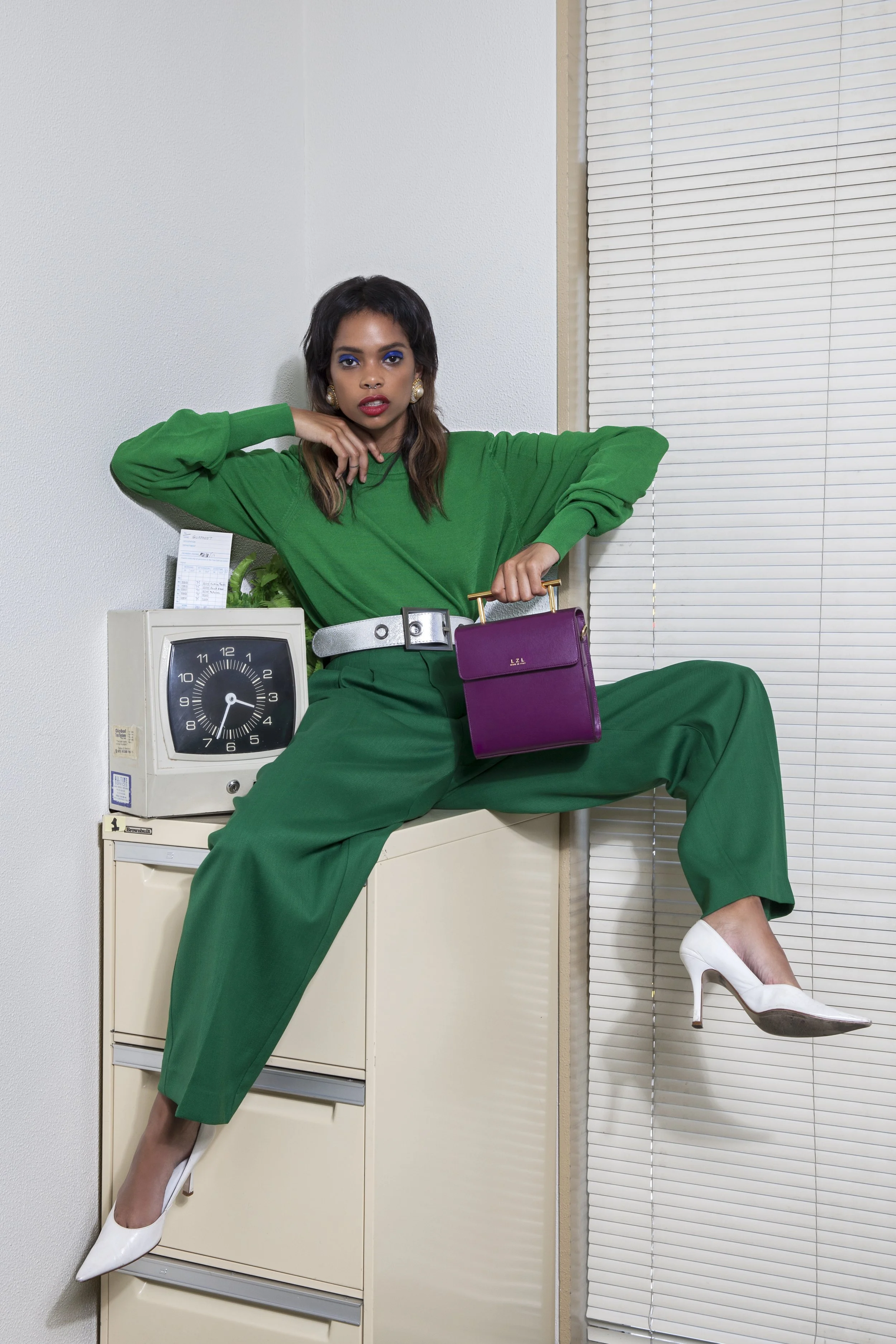 Woman in a green suit with white heels, sitting on a filing cabinet, holding a purple handbag, posing with one leg extended and one arm resting on her head, background featuring a white wall and closed blinds.