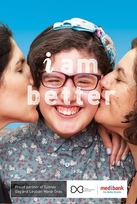A young girl with glasses and a floral shirt smiling as she is kissed on each cheek by two women, with a blue sky background. Overlaid text reads 'i am better'.