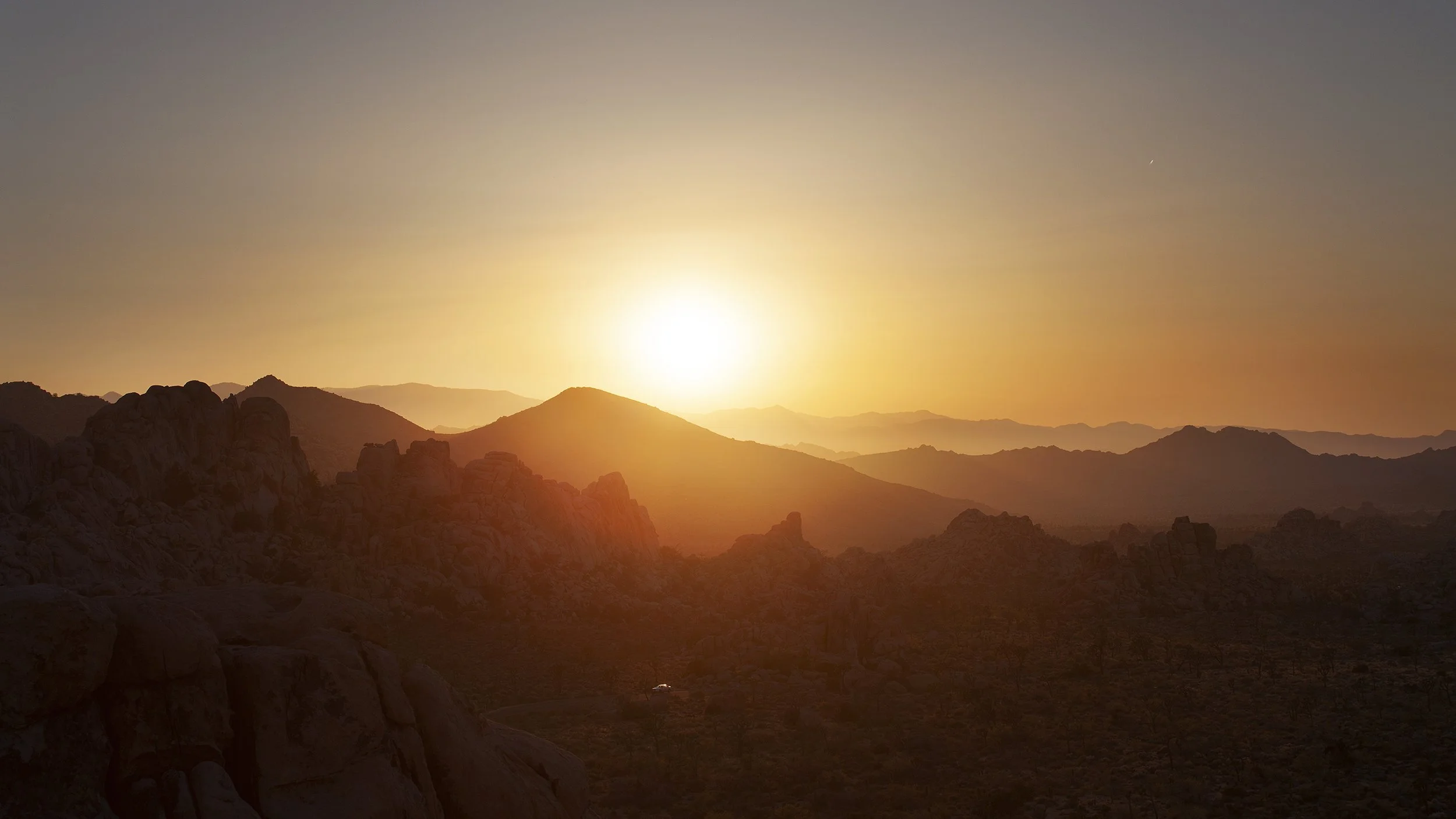 Sunset over a mountain range with rocky peaks and layered hills in the distance.