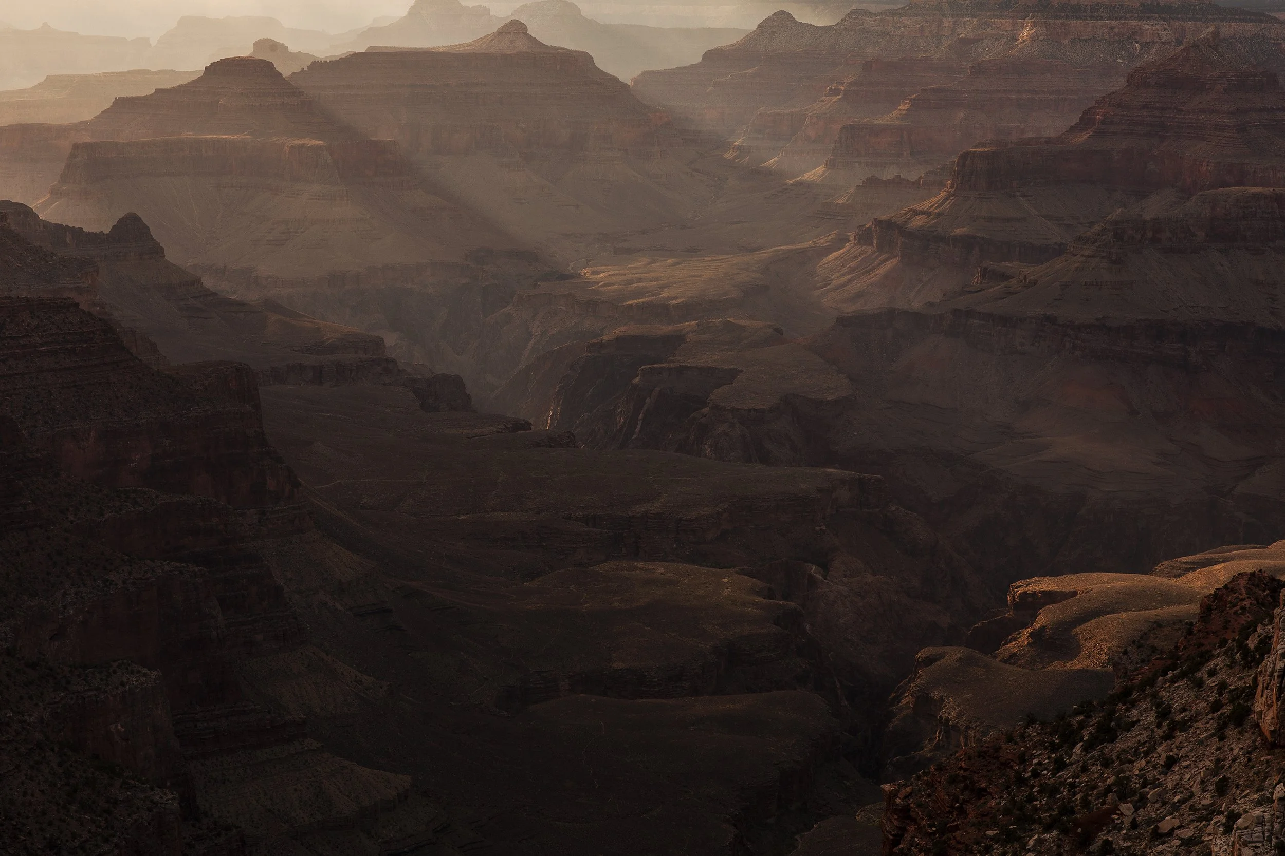 A wide view of the Grand Canyon displaying layered rock formations and deep valleys with shadows cast by the setting or rising sun.