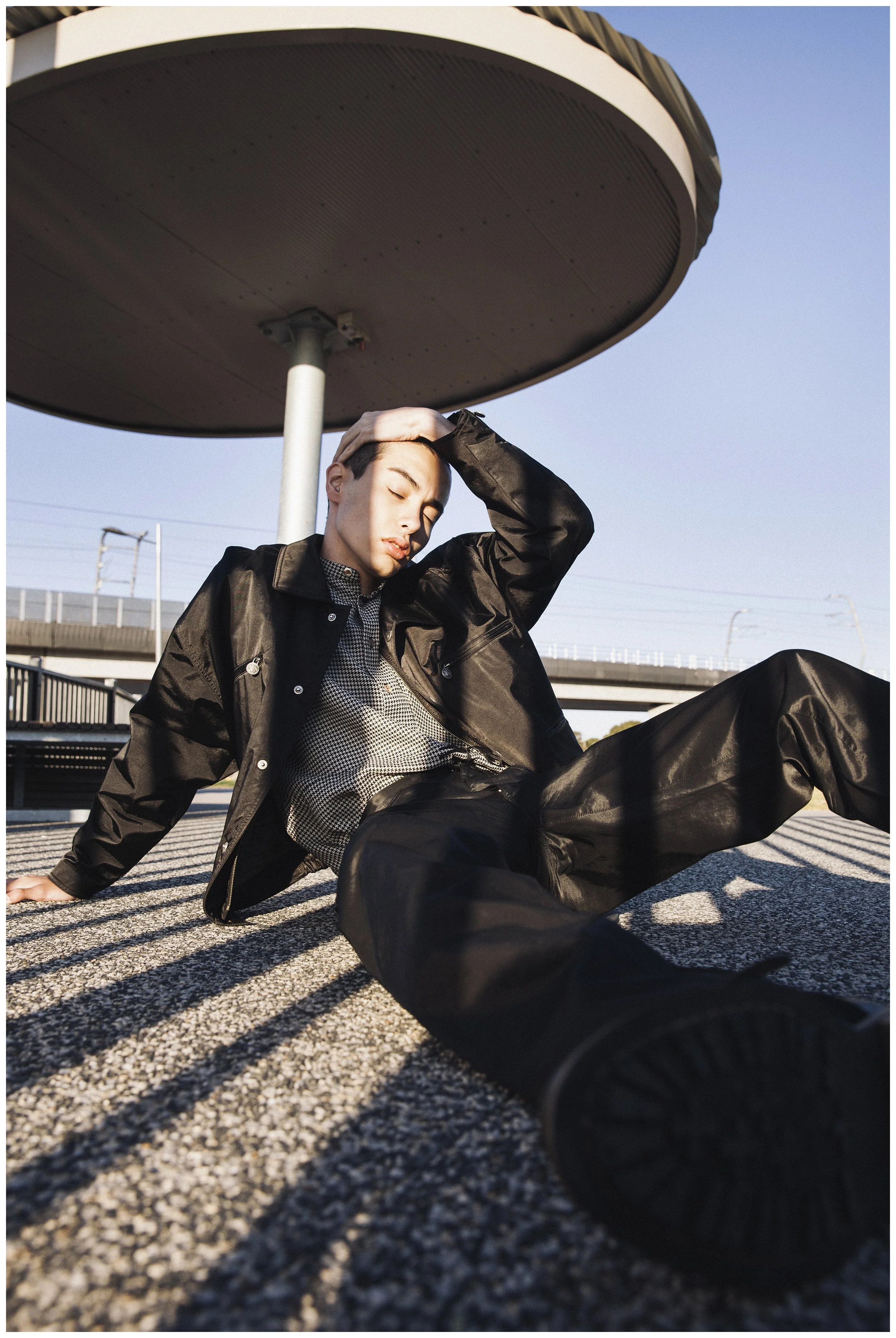 A young person with short hair in a black jacket and pants, sitting on the ground under a large, round, elevated structure, with one hand on their head and eyes closed, during sunset.