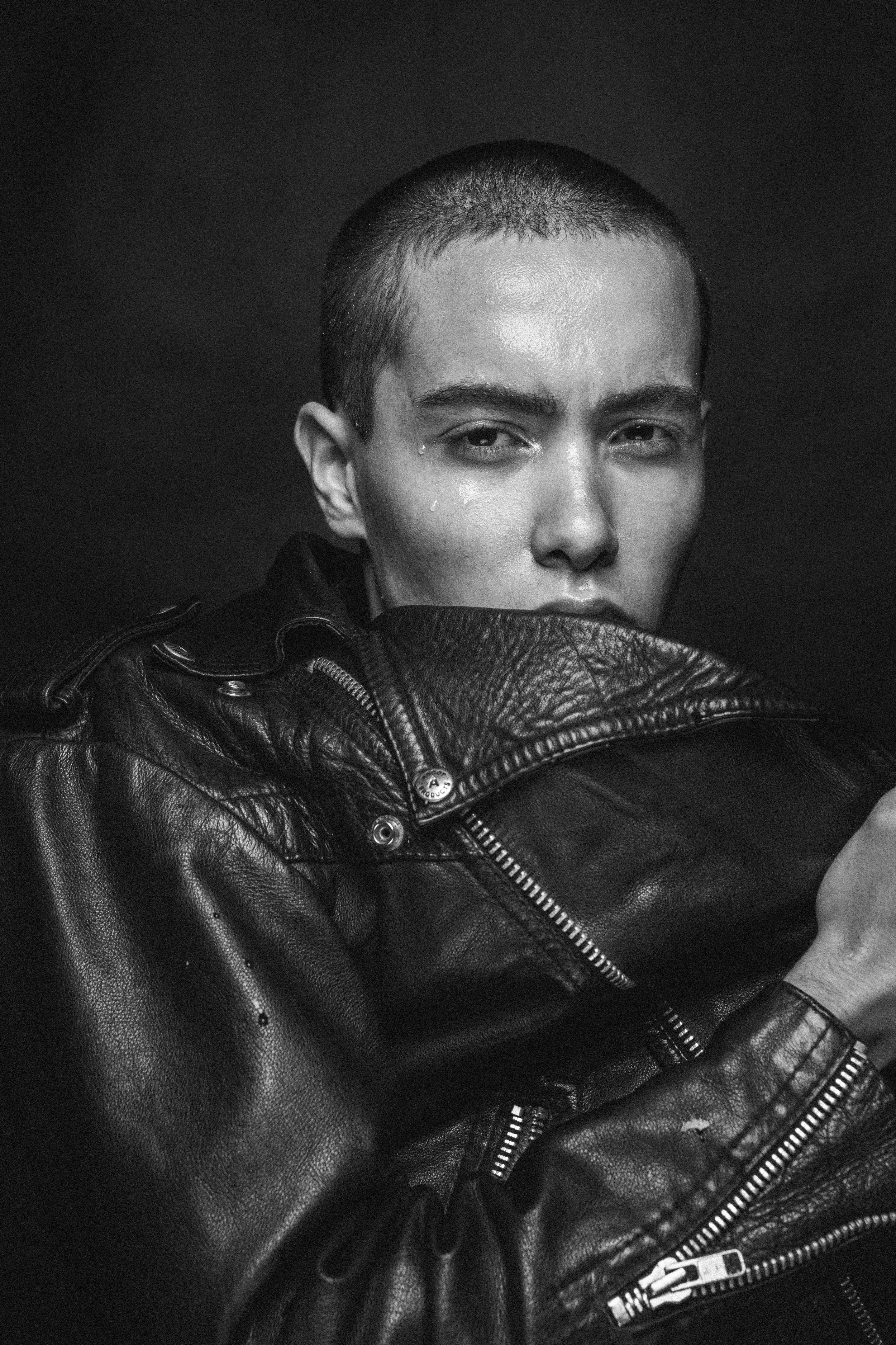 Black and white portrait of a person with short hair, wearing a leather jacket, looking directly at the camera with a serious expression.