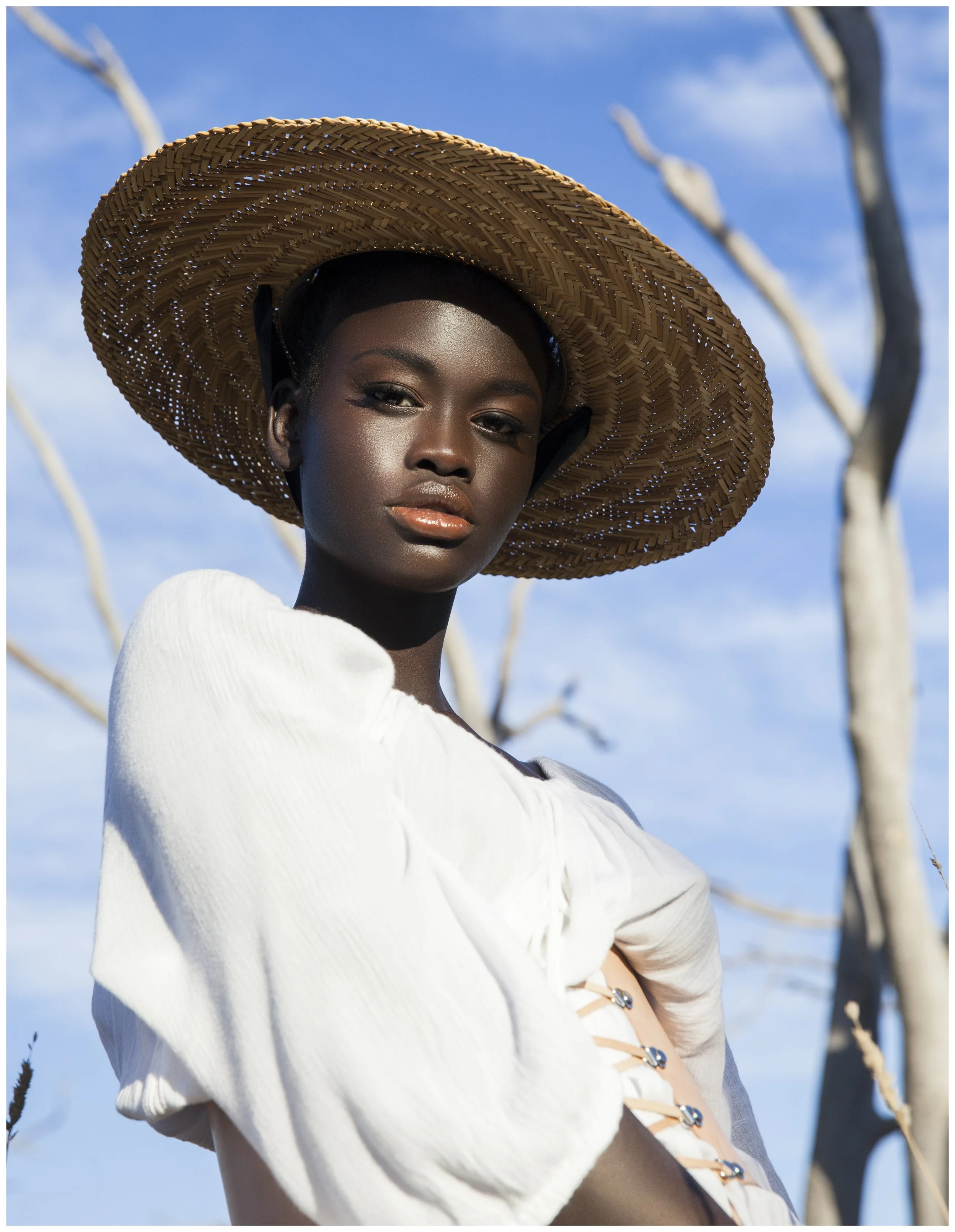A woman with dark skin, wearing a large woven straw hat and a white blouse, standing outdoors against a background of blue sky and leafless trees.