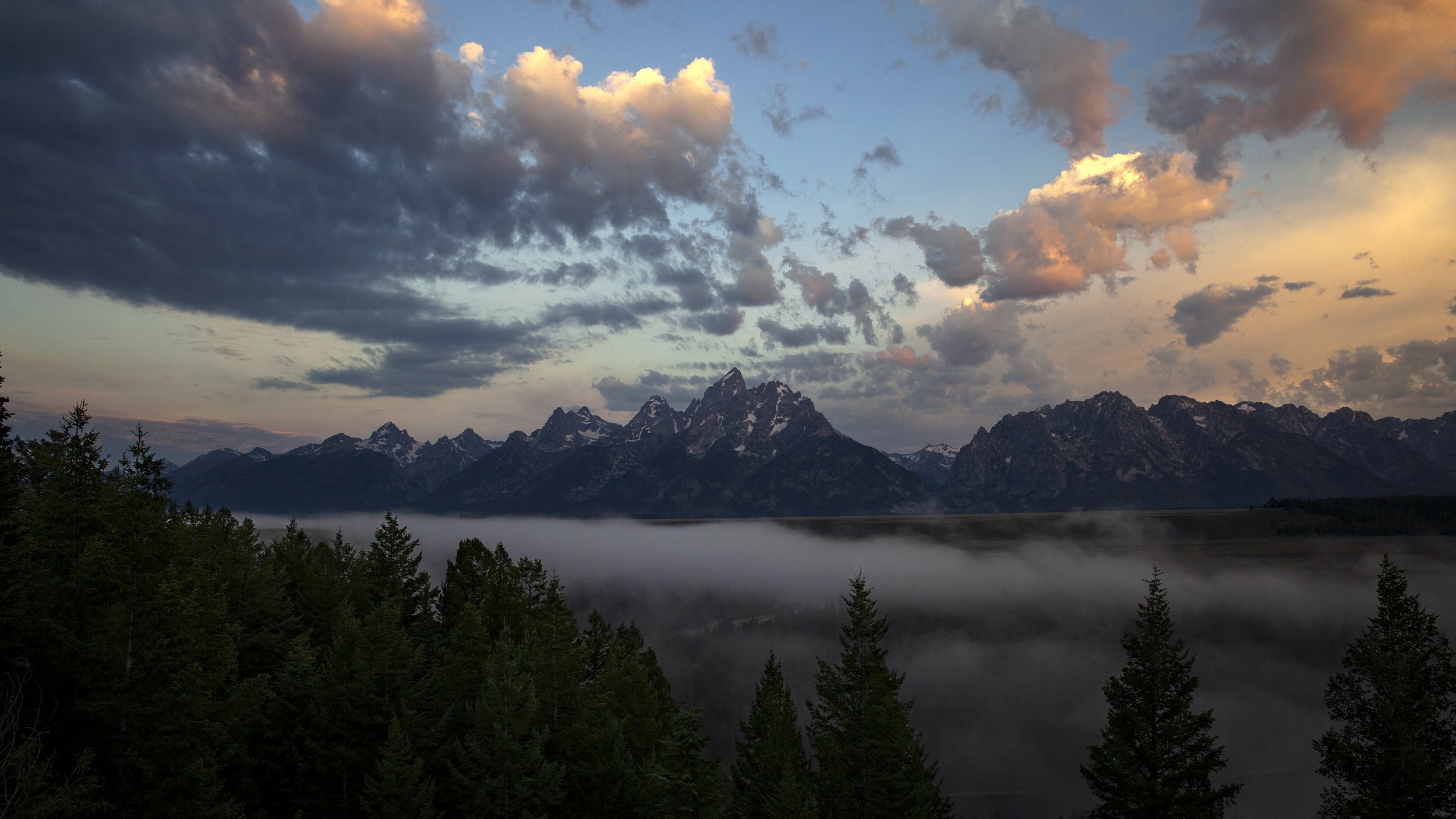 Mountain range with steep, snow-capped peaks under a cloudy sky during sunset, with a forest in the foreground and mist rising from a valley.