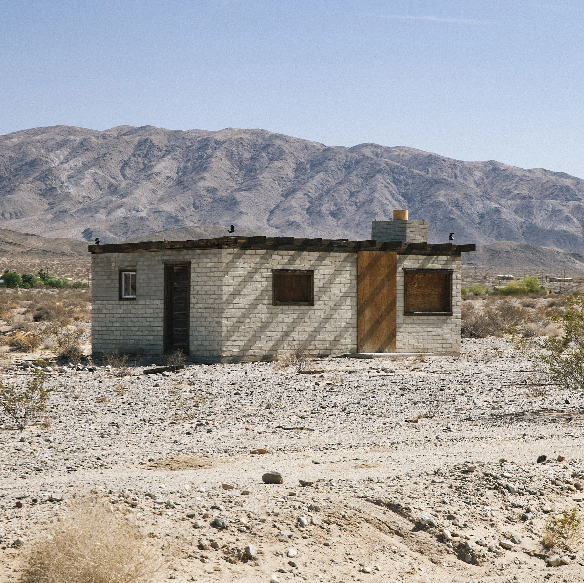 A small, abandoned brick house with boarded-up windows, set in a desert landscape with mountains in the background.