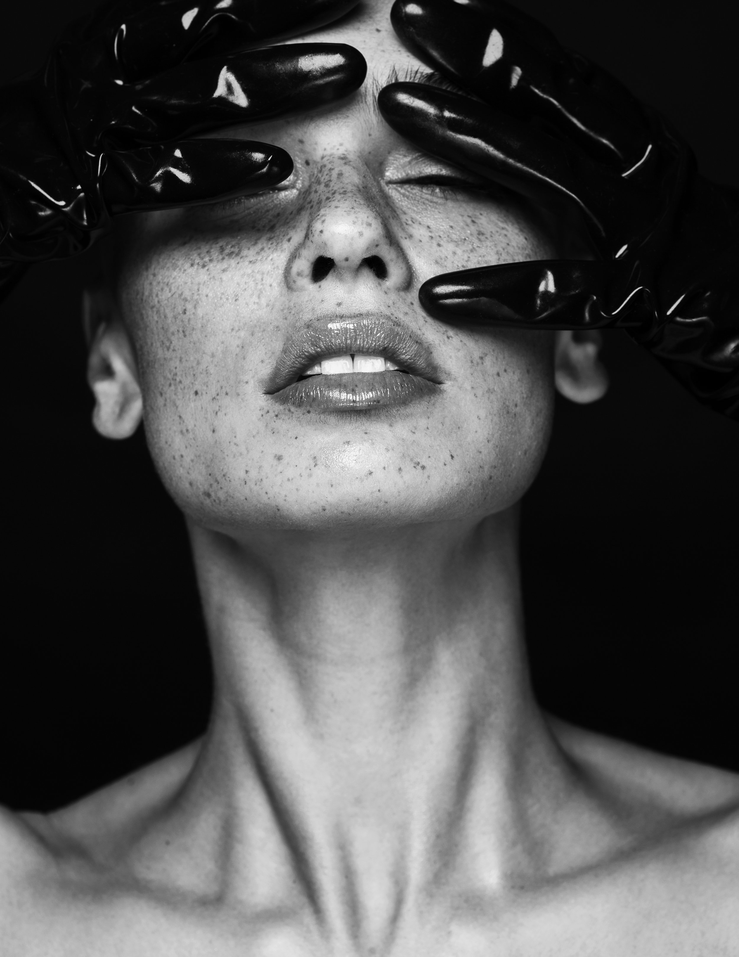 Close-up black and white photo of a woman with freckles, lips slightly parted, eyes closed, with hands in black gloves touching her face.