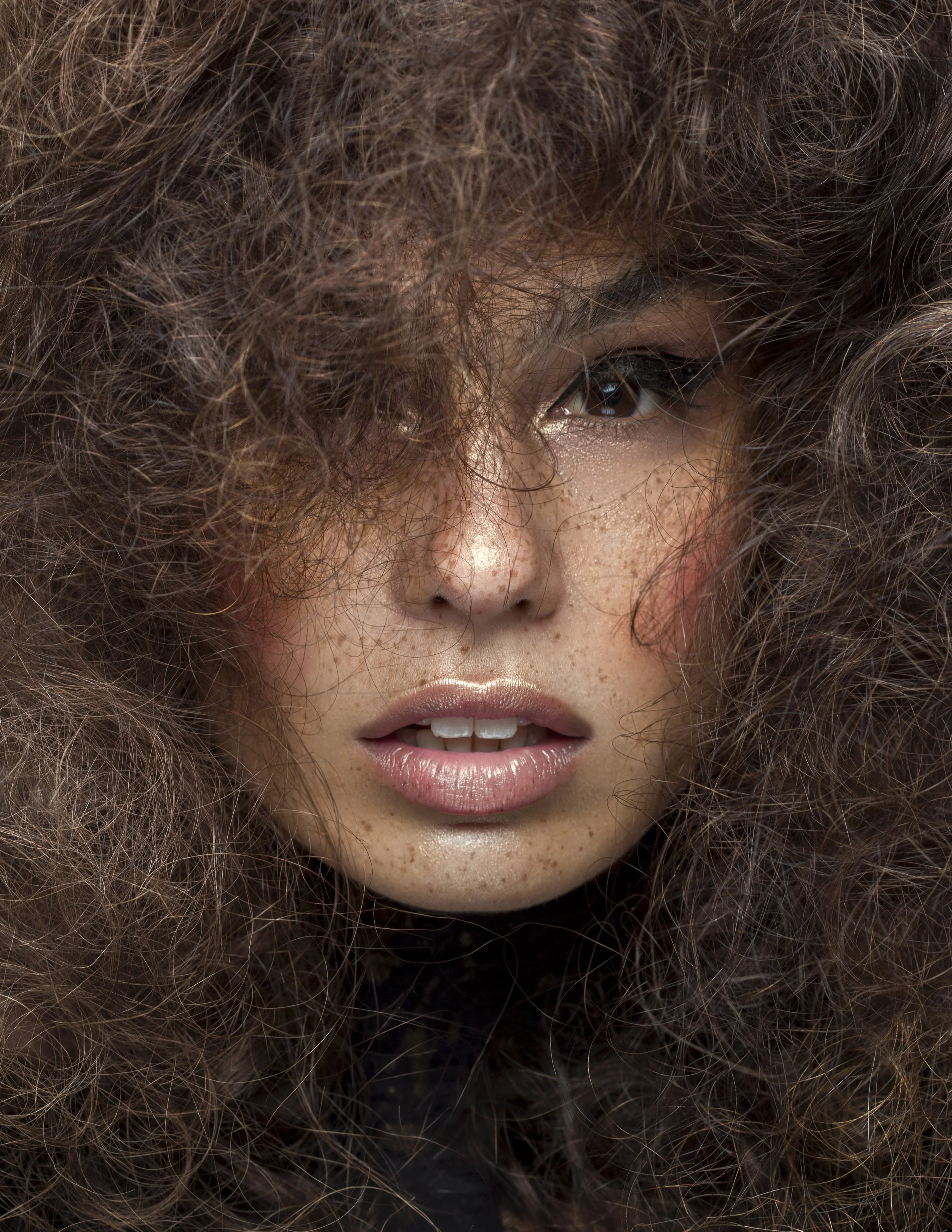 Close-up of a woman with voluminous, curly brown hair partially covering her face, with freckles, shiny lips, and makeup accentuating her eyes.