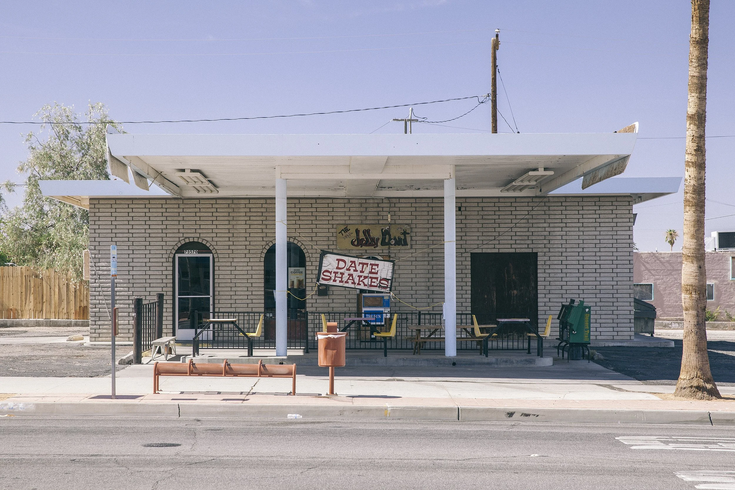 A small cafe with a sign that reads 'The Jelly Donut' and a larger sign advertising 'Date Shakes.' The building has a brick facade, chairs and tables outside, and a trash can. There is a sidewalk in front with a street and a parking meter.