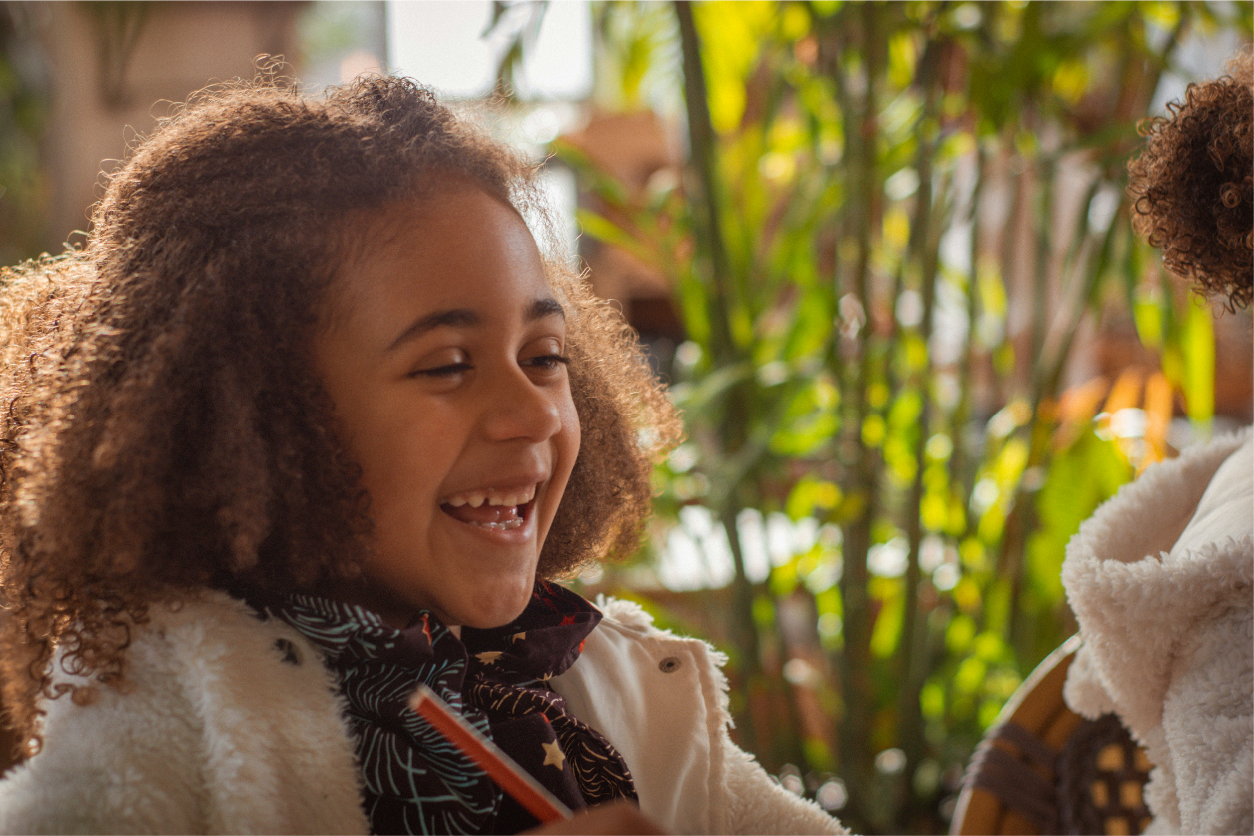 A smiling young child with curly hair sits indoors in warm sunlight, holding a pencil and looking toward another person, with green plants in the background.
