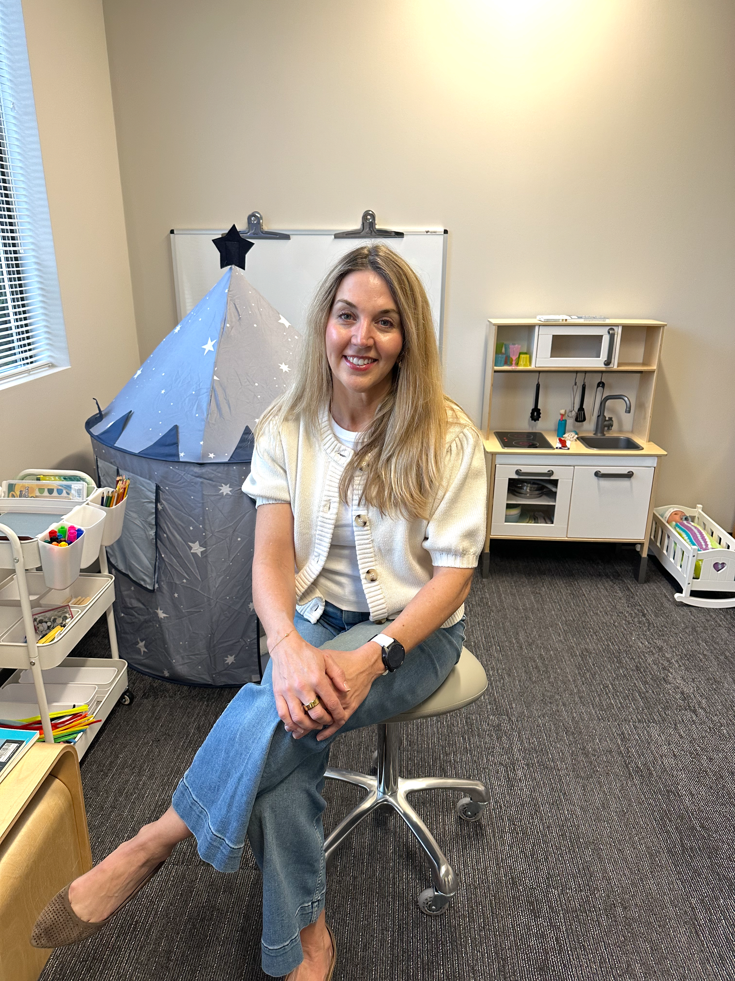 A woman with long blonde hair, wearing a cream-colored cardigan and jeans, sitting on a stool in a playroom or child's room. She is smiling, with her left foot on the floor and her right ankle resting on her left knee, showing a brown sock and tan shoe. Behind her, there is a small play tent with a star pattern, a white toy kitchen set, and various toys and art supplies in containers.