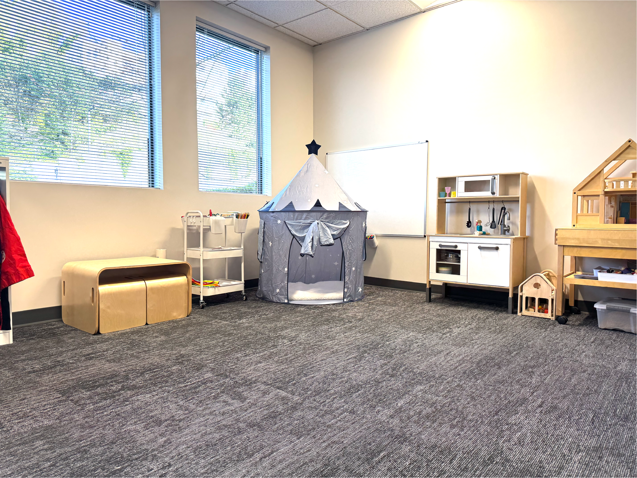 A room with large windows, a play tent shaped like a cone with stars and a moon, a small play kitchen set, a wooden dollhouse, and a plastic container on the floor.