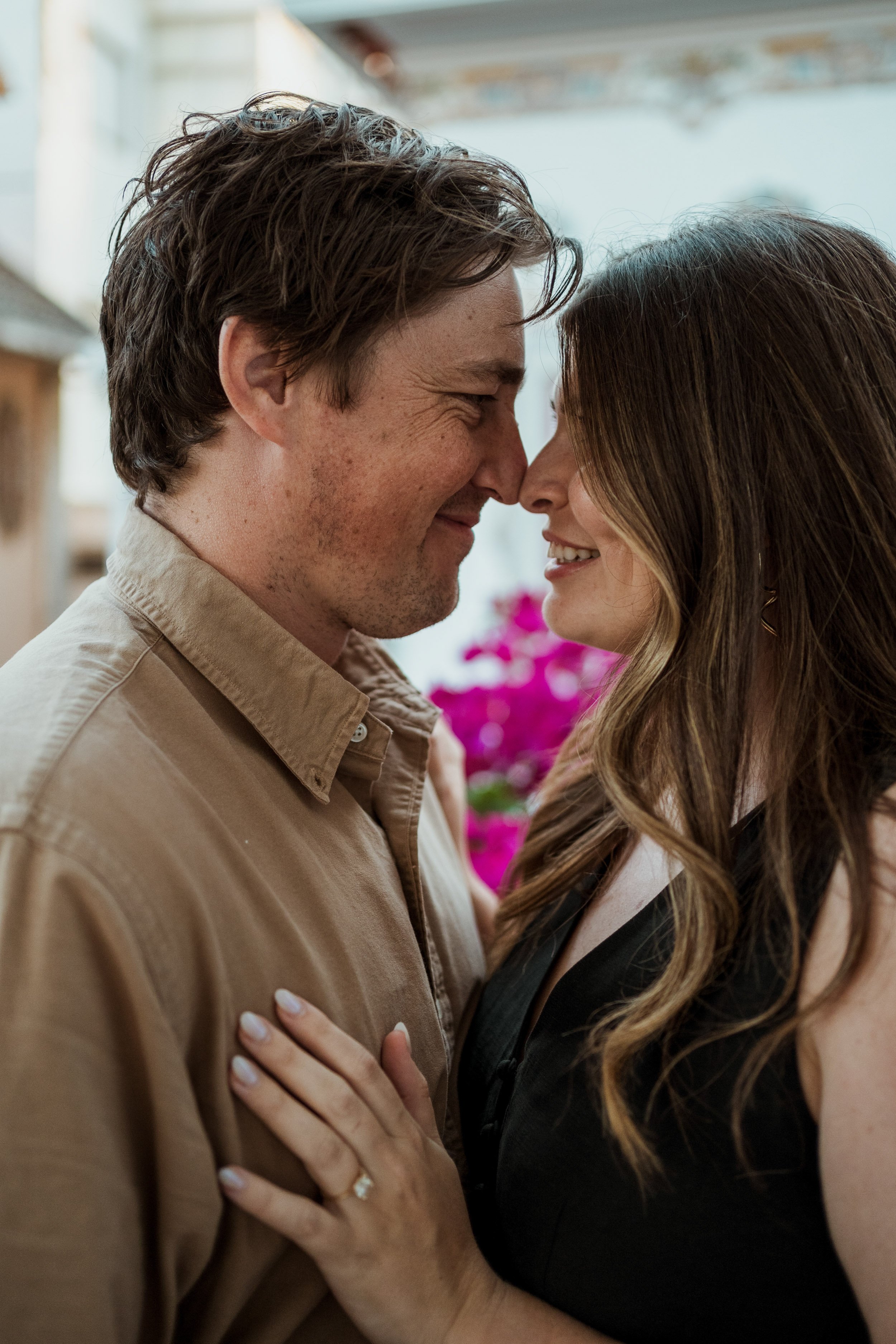 A couple with their noses touching, smiling, holding each other close, outdoors with pink flowers in the background.