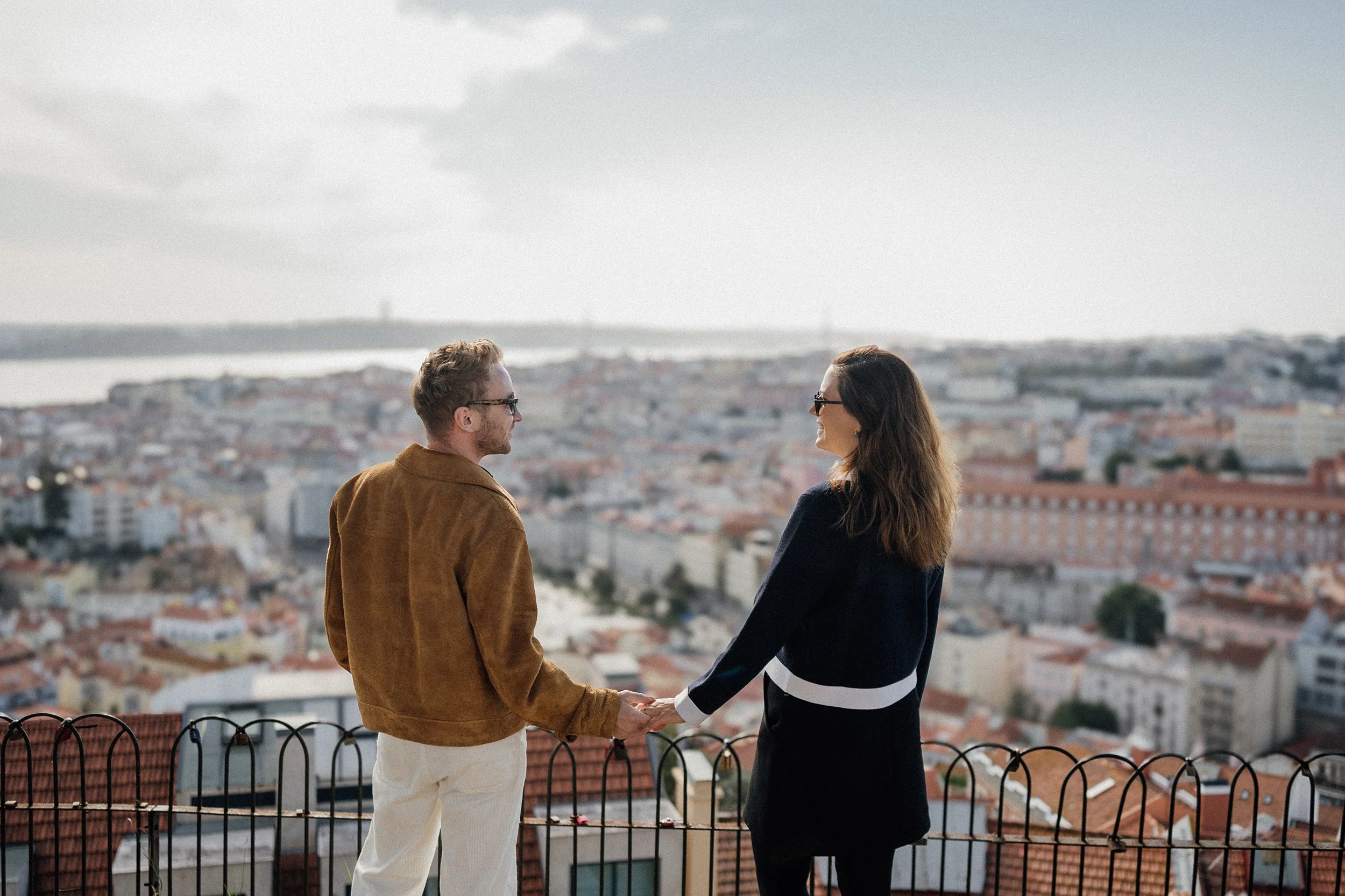 Engagement photography in Lisbon, couple holding hands at scenic city viewpoint overlooking historic rooftops.