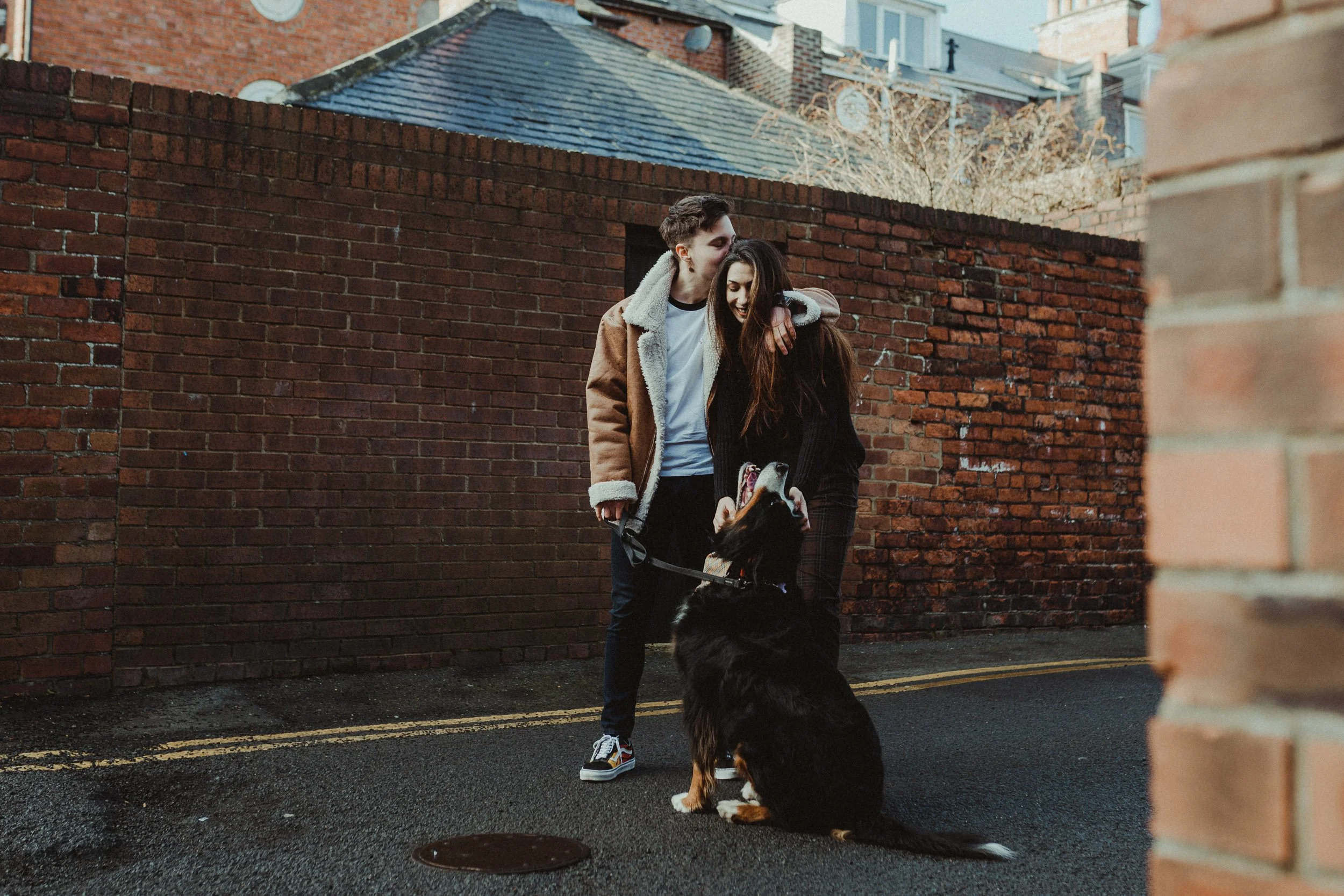 Candid engagement photography in London, couple standing together against brick wall in urban setting with dog.