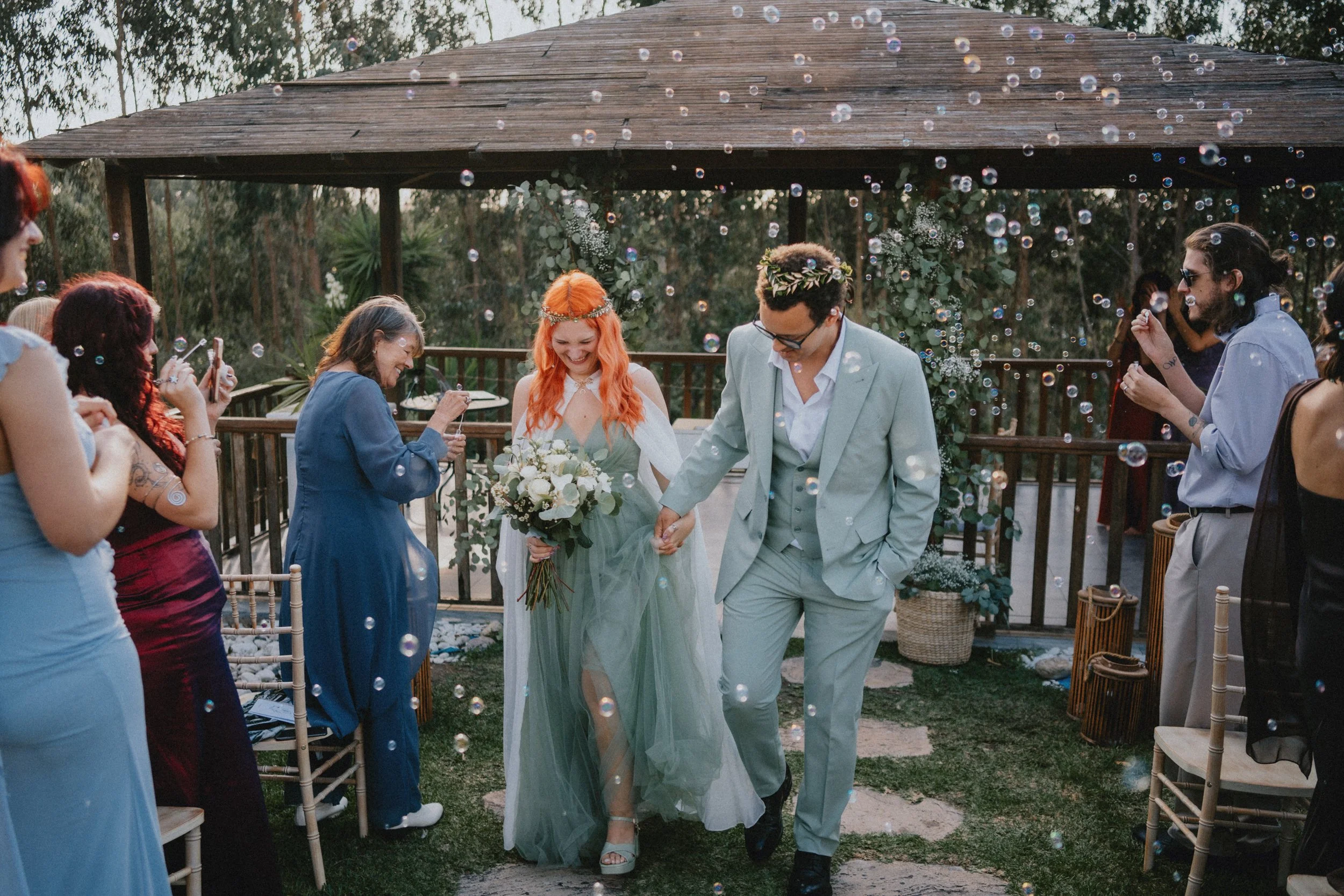 Outdoor garden wedding ceremony in Portugal with alternative couple walking down the aisle surrounded by guests and water bubbles