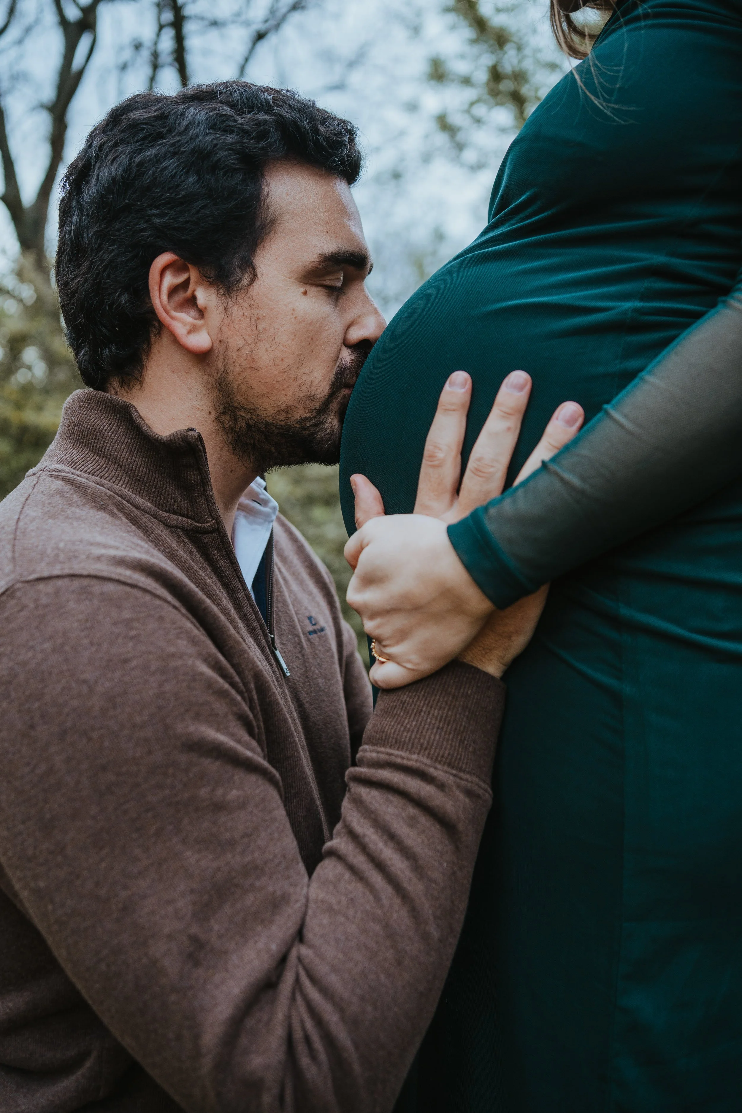 Intimate pregnancy photography of partner kissing baby bump during outdoor maternity session.