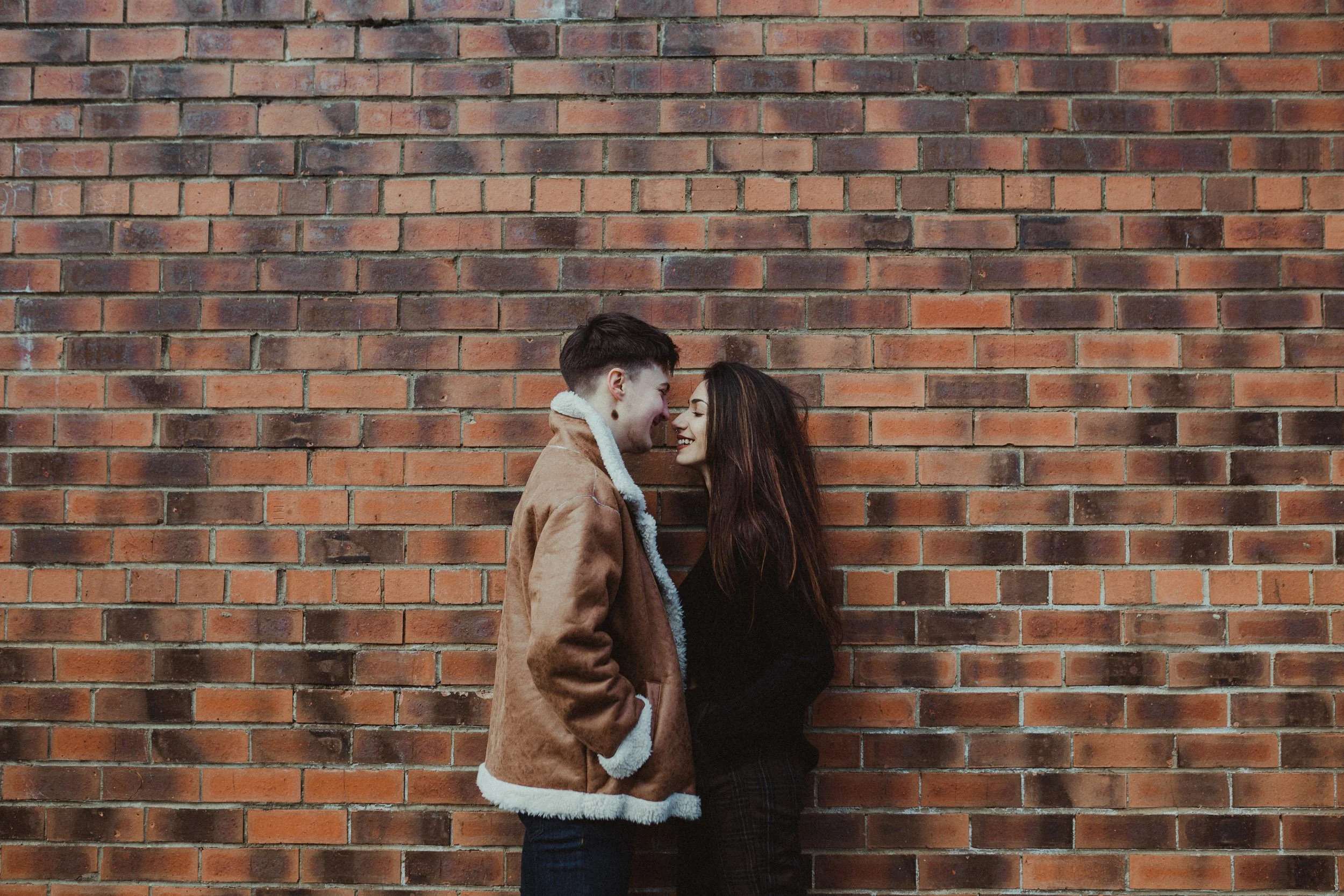 Candid engagement photography in London, couple standing together against brick wall in urban setting.