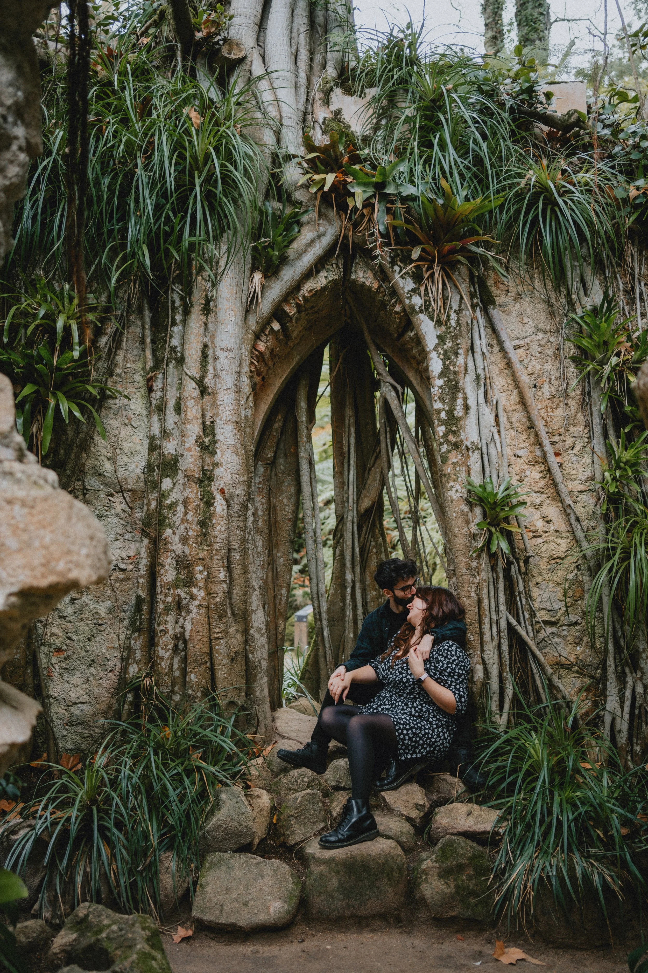 Engagement photography in Sintra forest, couple standing beneath natural stone arch surrounded by greenery.