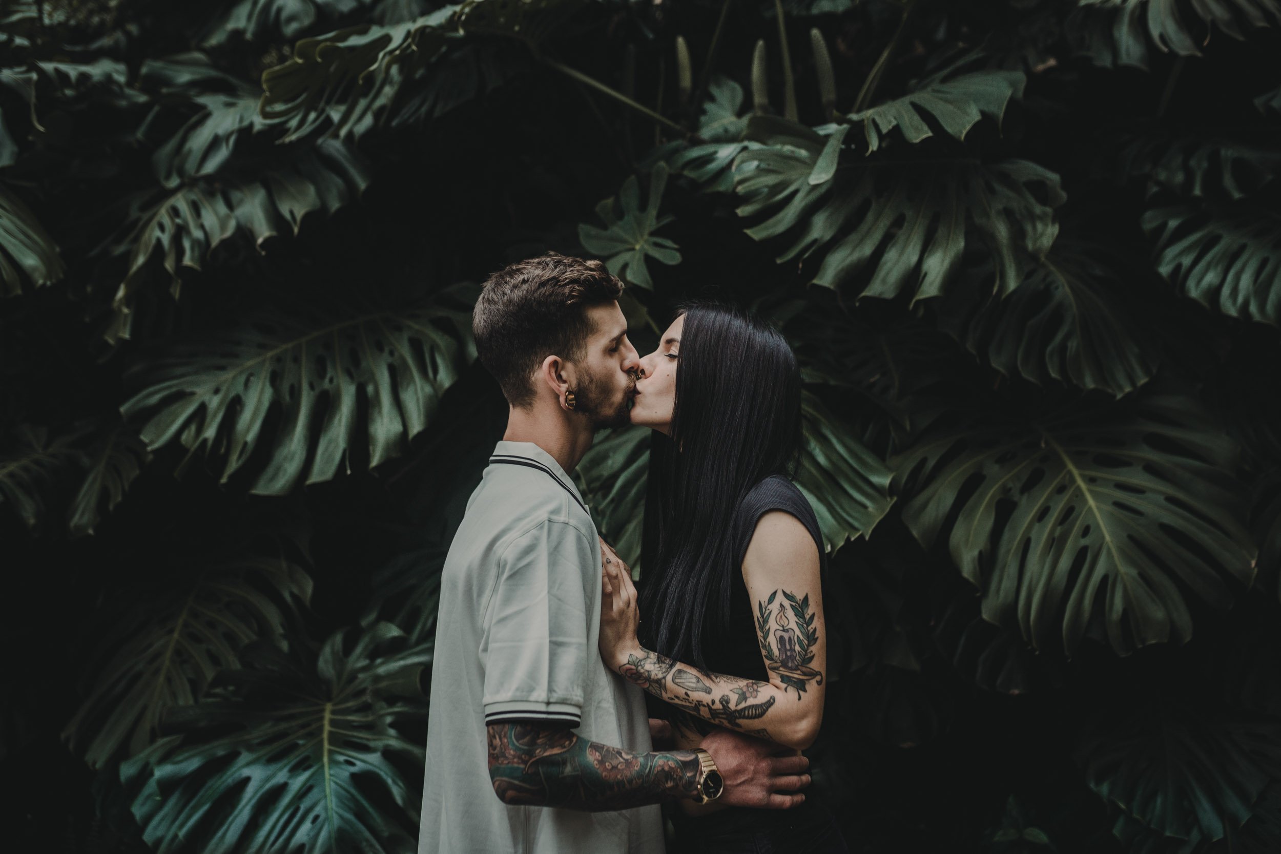 A couple sharing a kiss in front of lush green tropical leaves.