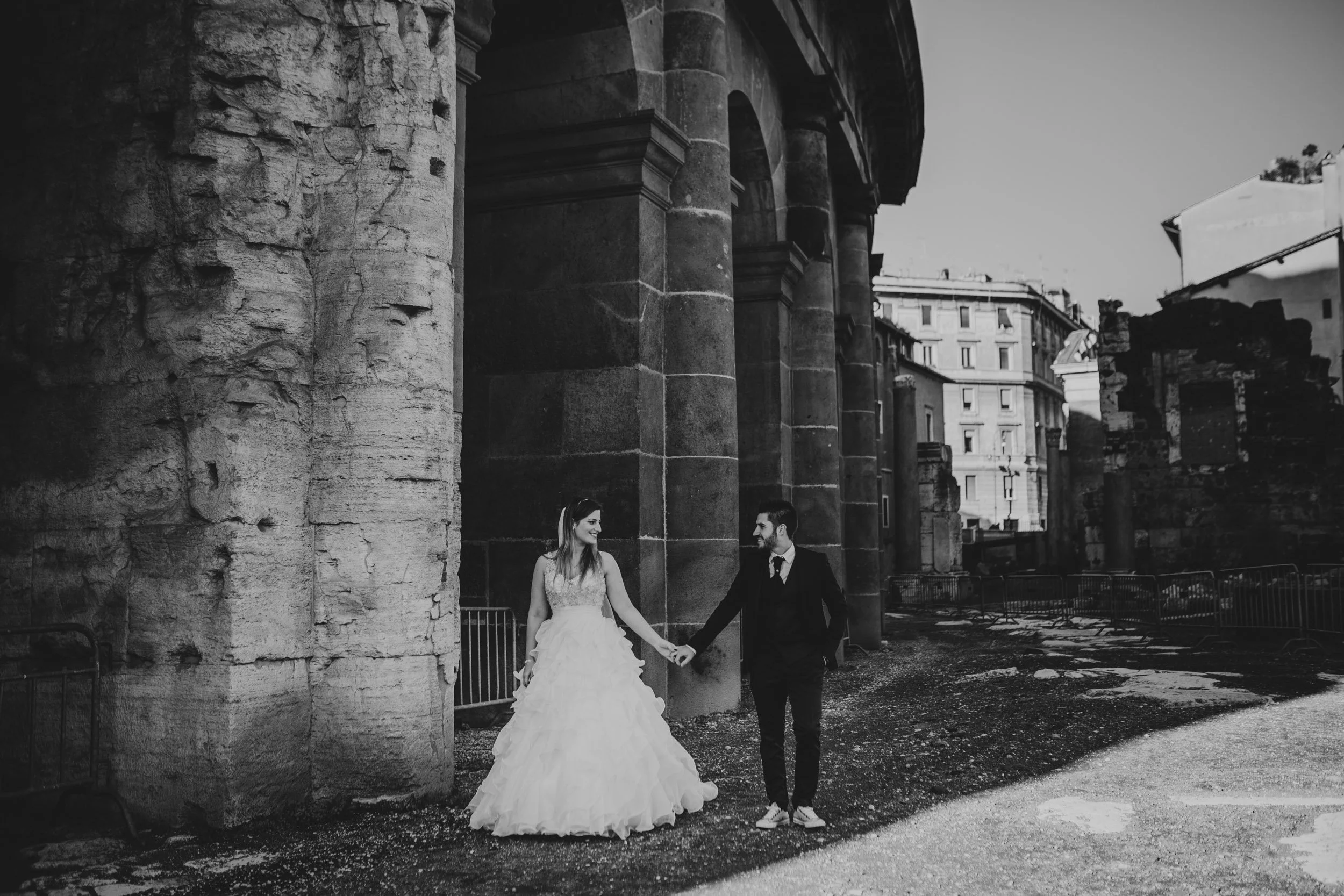 Black and white photo of bride and groom holding hands under historic Roman columns