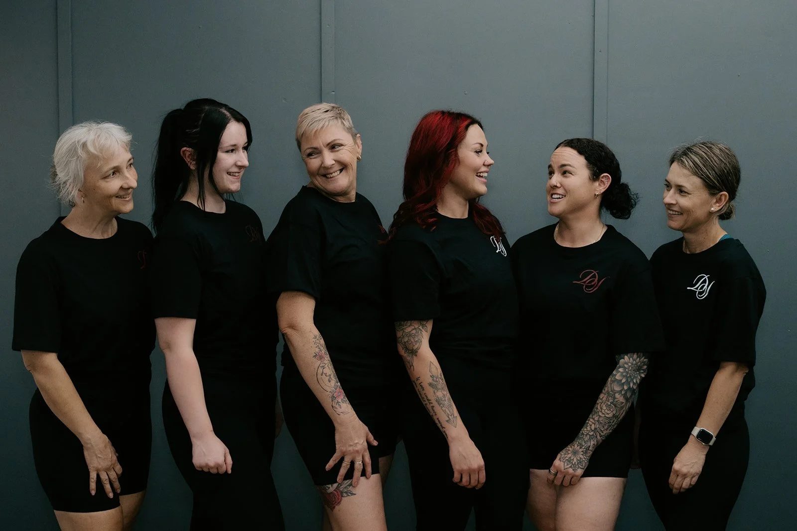 Six women standing in a line against a gray wall, smiling and interacting with each other.