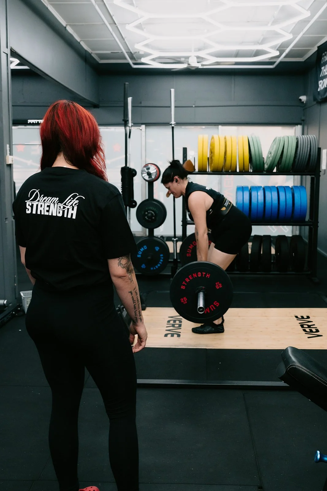 A woman with red hair standing and observing another woman lifting a barbell in a gym.