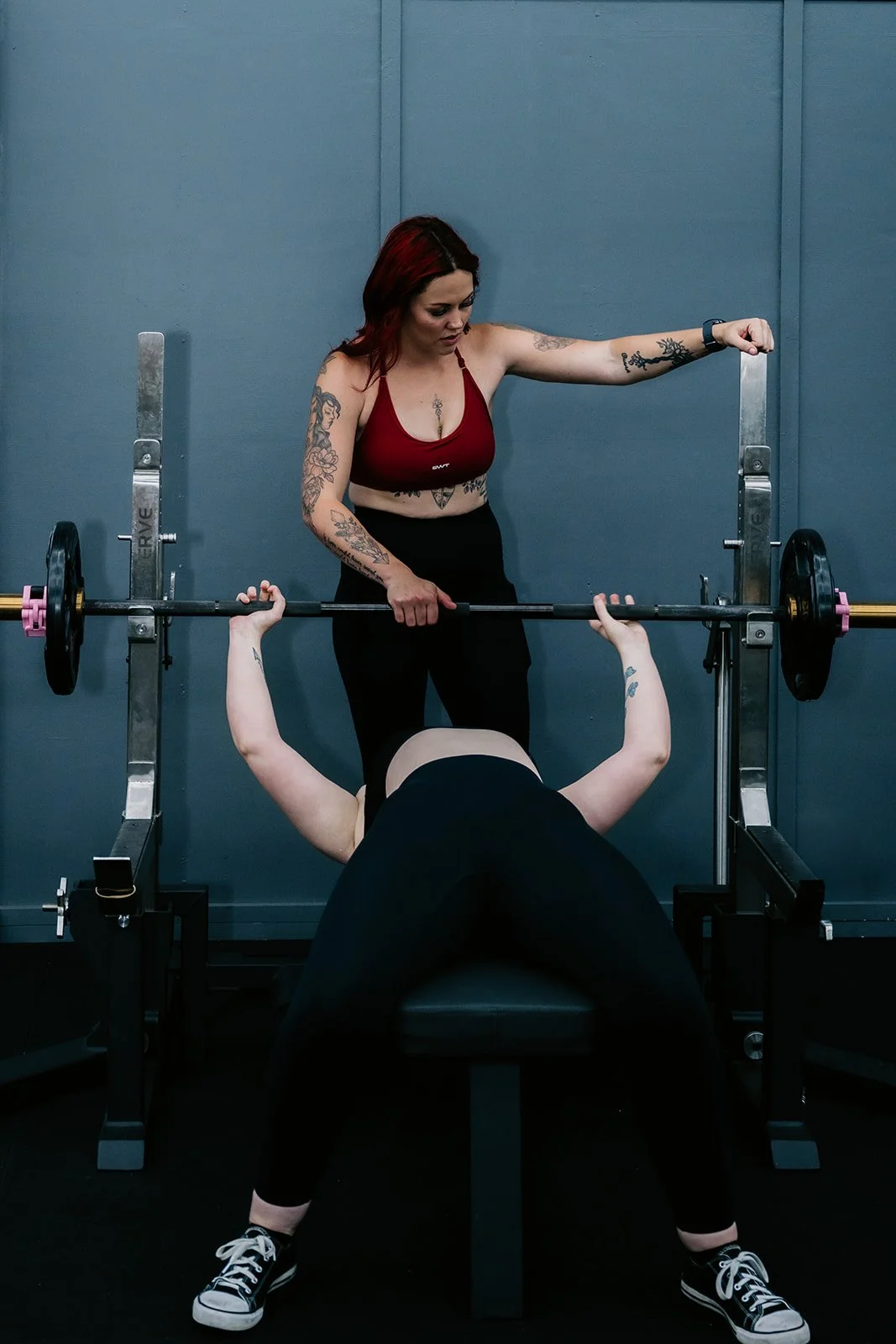 A woman in a red sports bra and black leggings assists another woman lifting a barbell on a bench press machine in a gym.